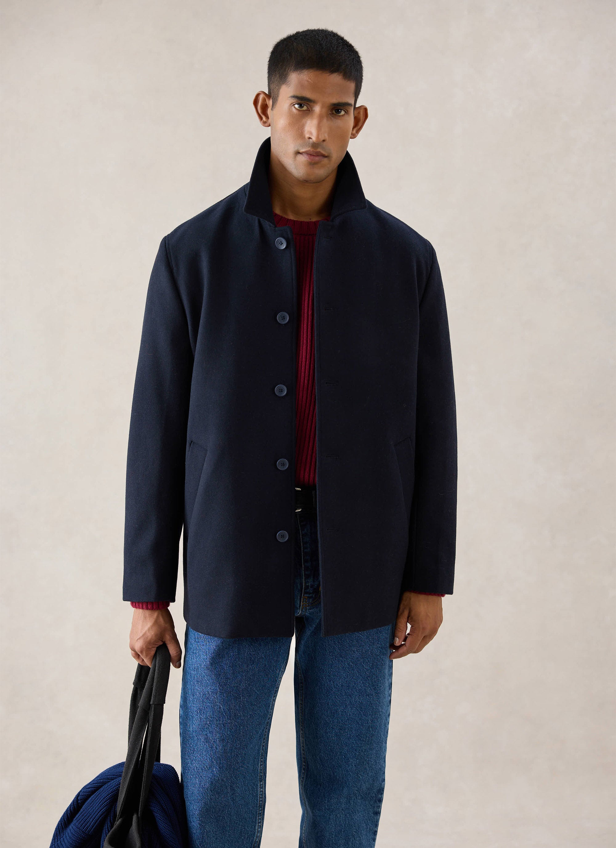 A model wearing the navy wool Percival pea coat over a red jumper and blue jeans, holding a bag.