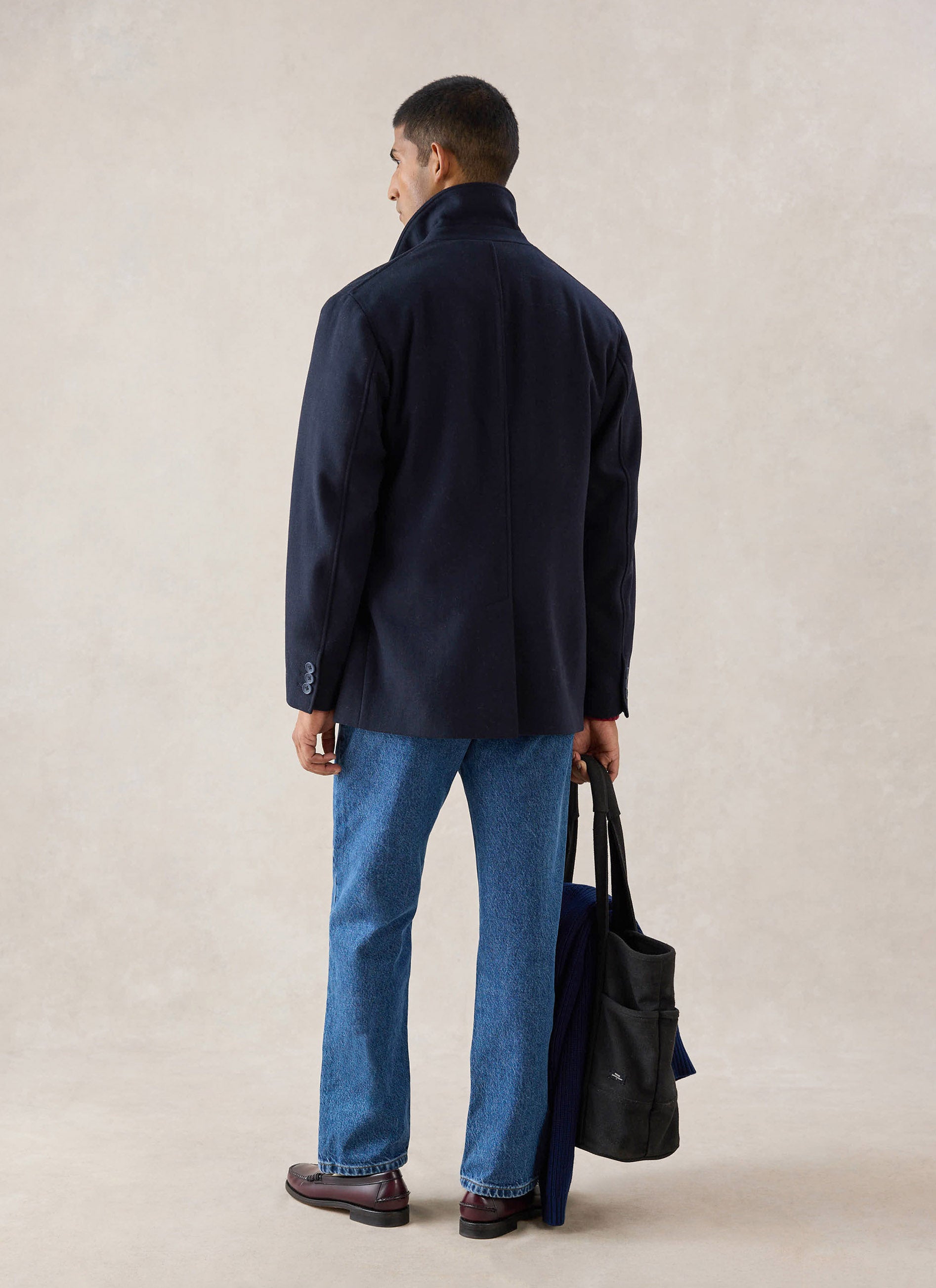 A model from the back wearing the navy wool Percival pea coat, styled with blue jeans and holding a dark tote bag.