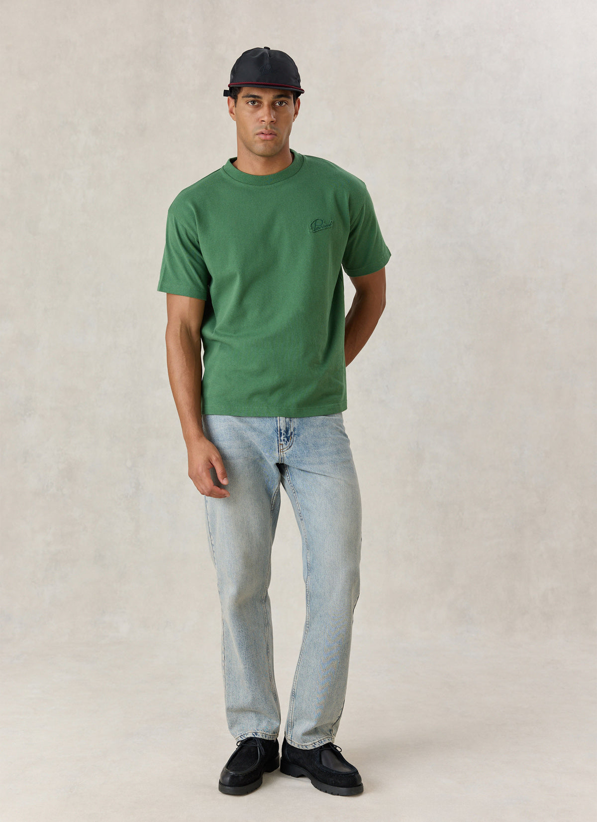A model wearing the Percival forest green organic cotton oversized t-shirt with light wash jeans and a black cap.