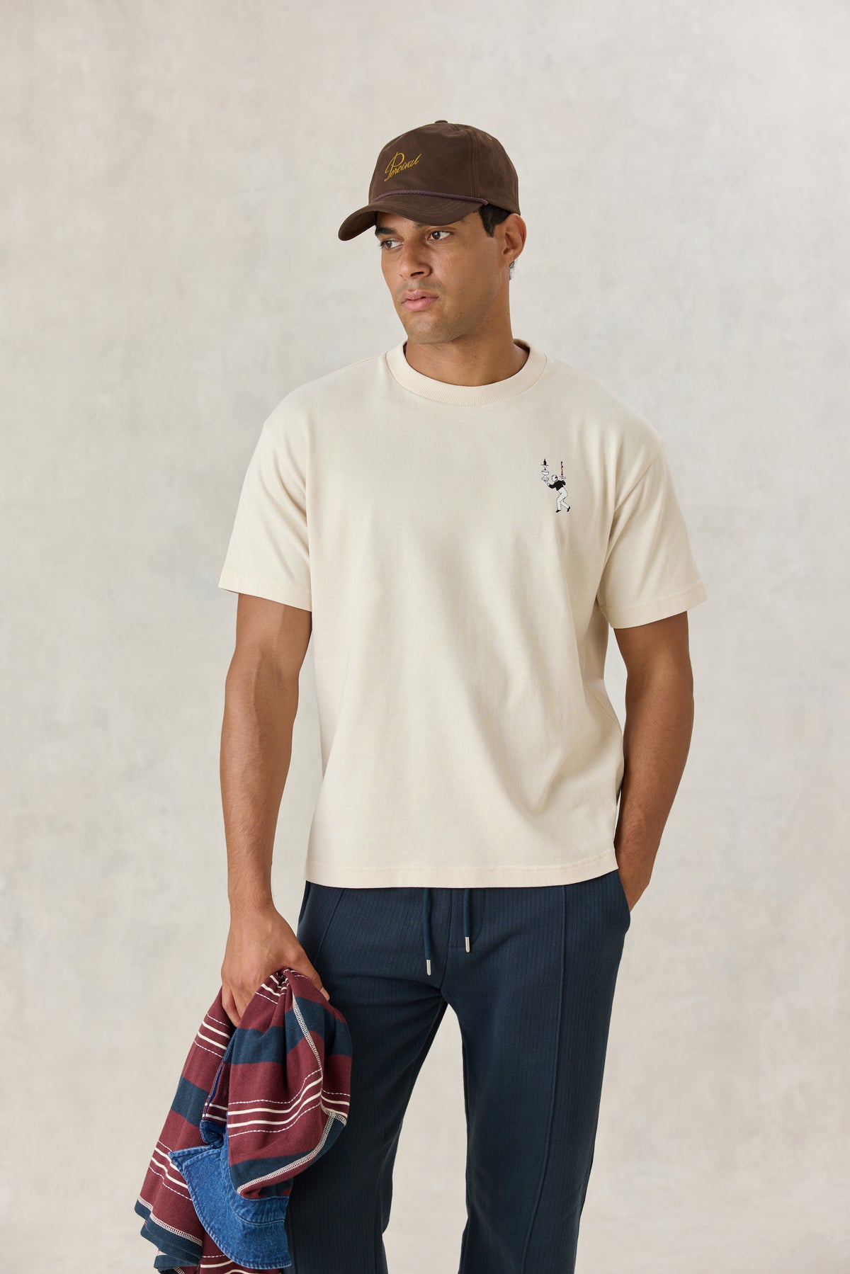 A model wearing the cream Percival Perci Waiter oversized T-shirt with a small chest graphic, brown cap, and navy trousers.