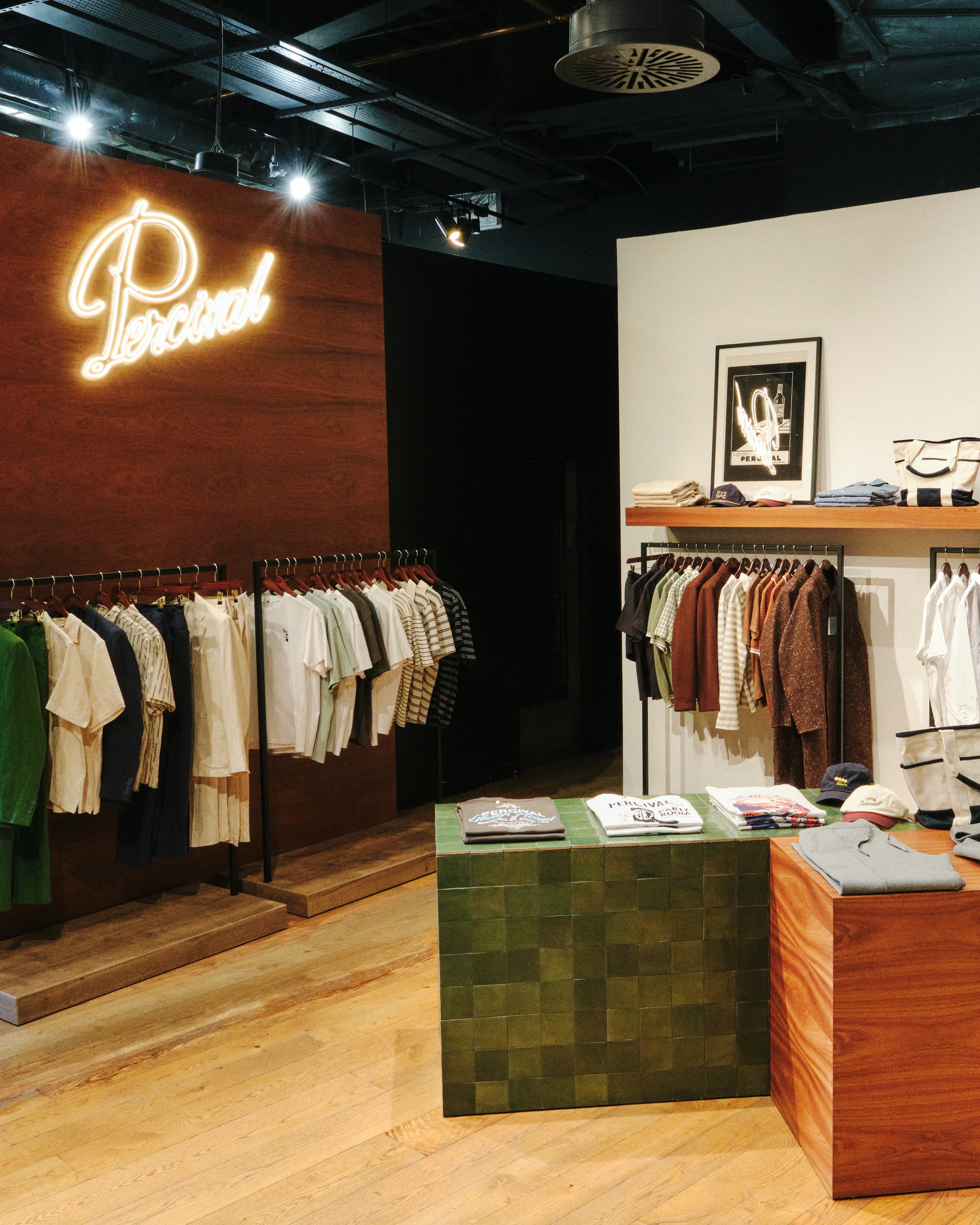 Glowing Percival neon brand sign on a wooden wall panel above rows of men's clothing racks at the Selfridges Birmingham pop-up store.
