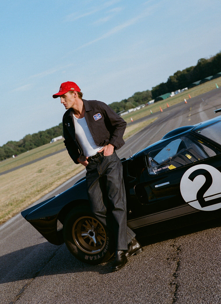 Model wears a navy Percival x Ford GT40 canvas jacket and straight leg jeans by a vintage race car on a racetrack.