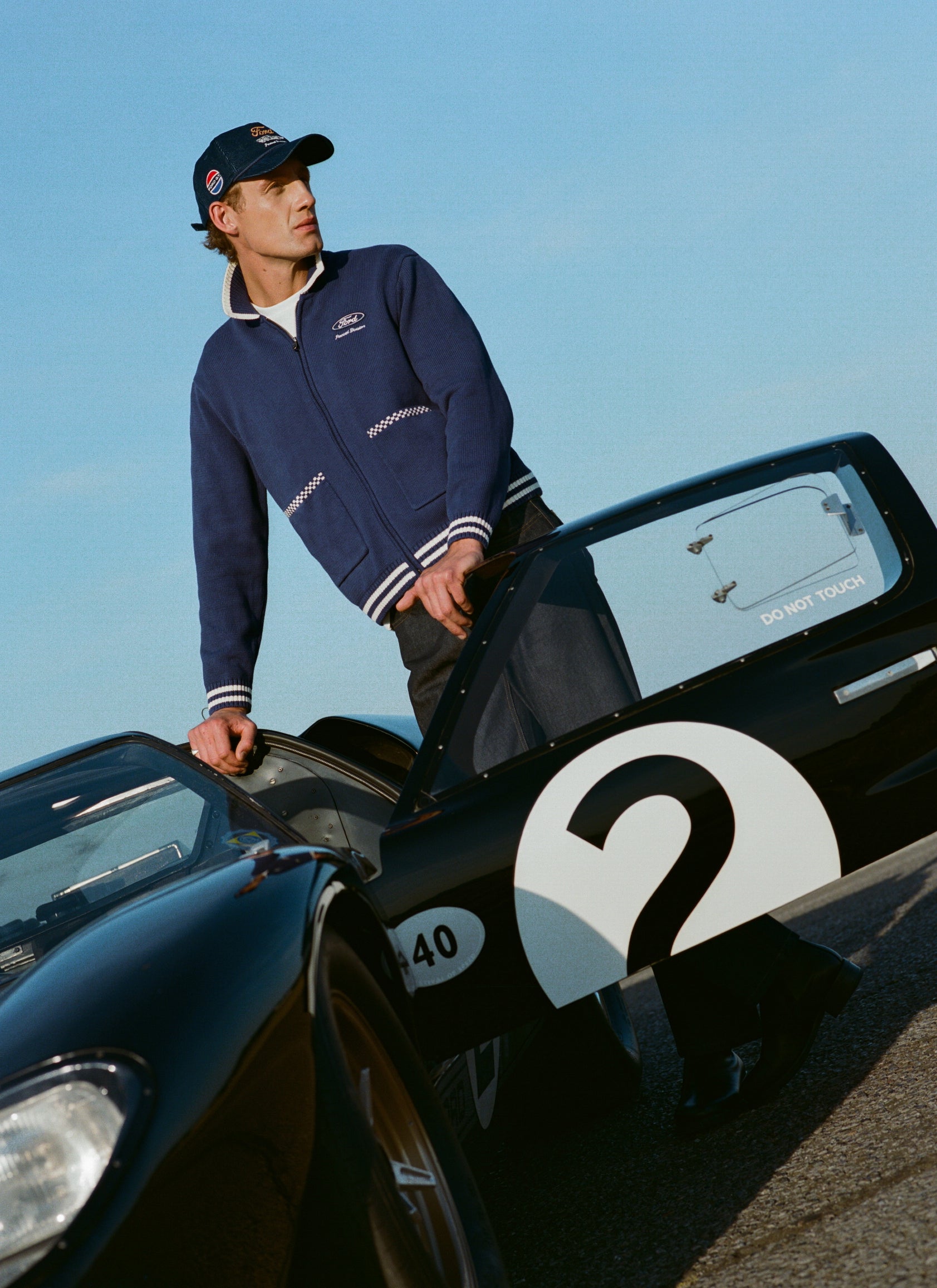 A model wears a blue Ford x Percival zip cardigan and navy cord cap, standing by a classic racing car outdoors.