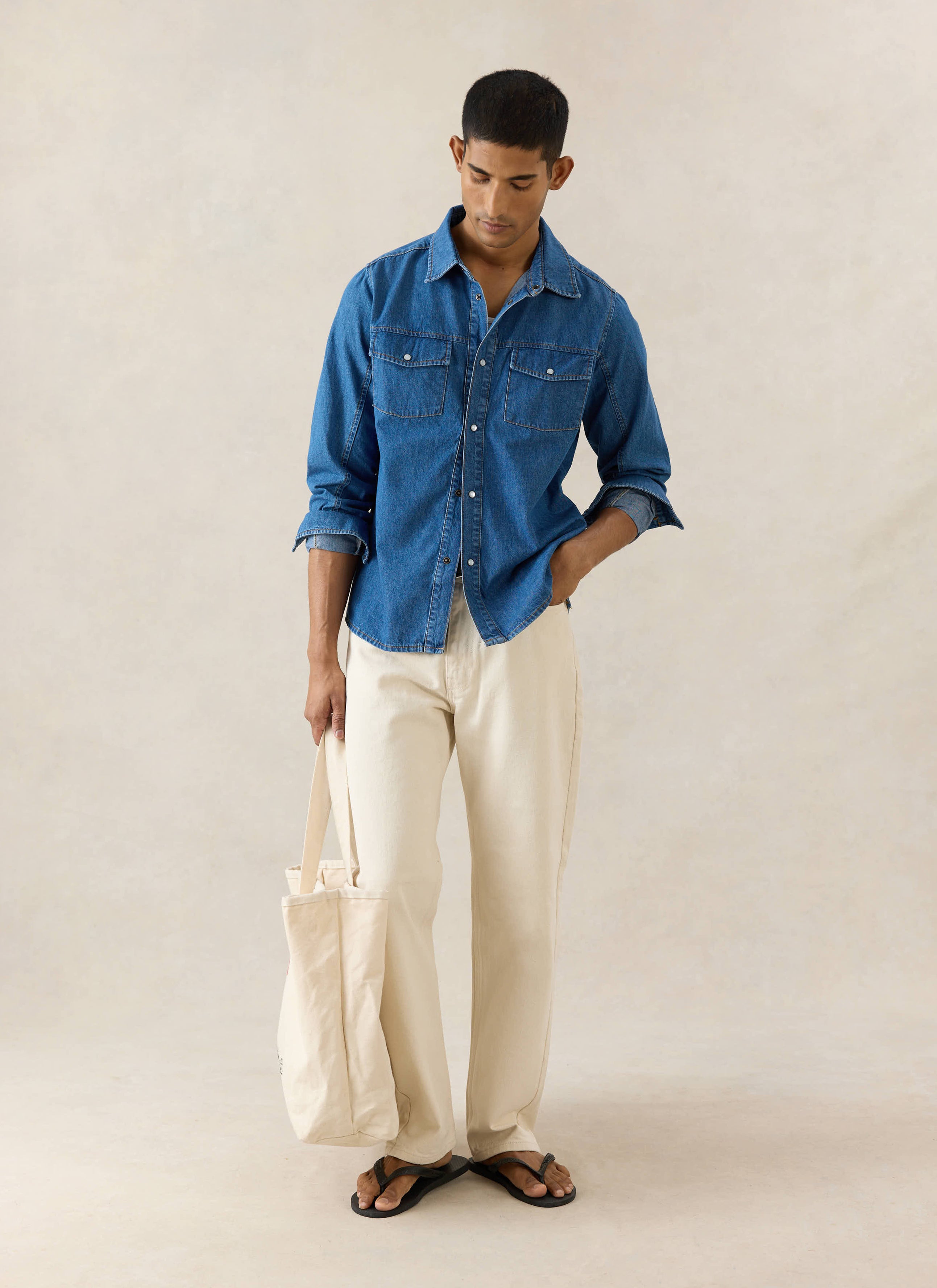 Mid wash blue Percival denim western shirt with pearl buttons, styled with cream trousers and a canvas tote bag.