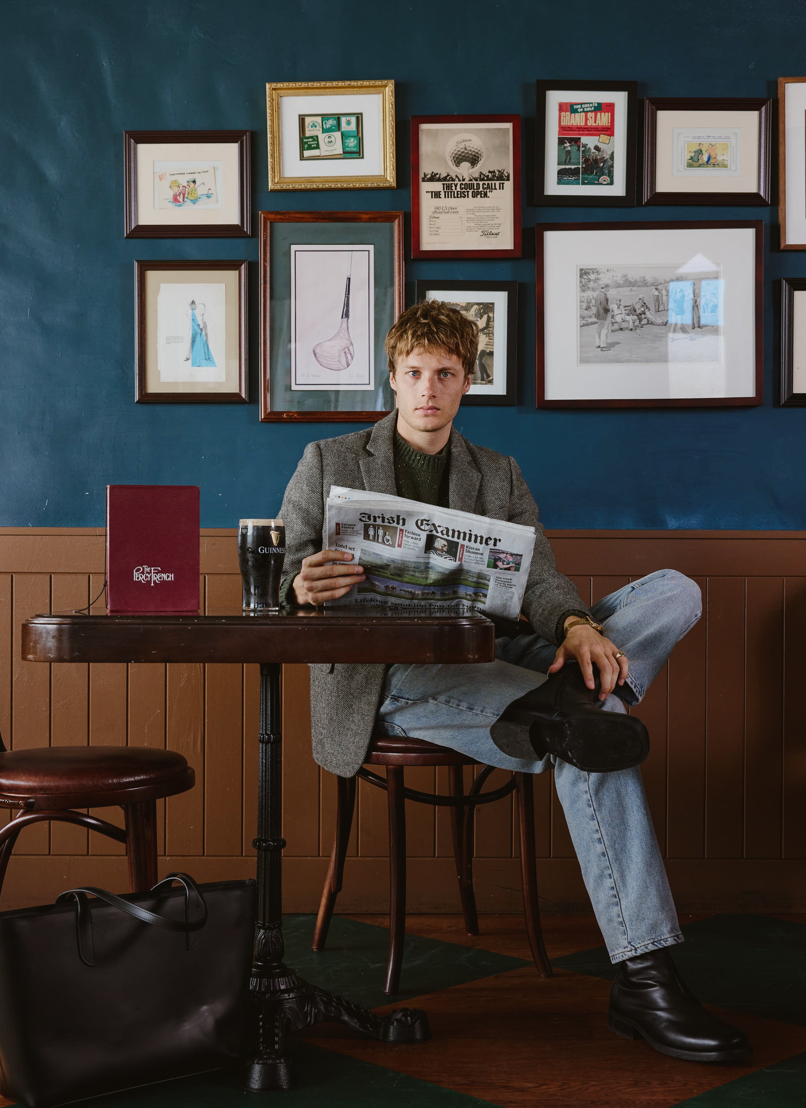 A model wears the Percival forest green Rhodes jumper under a patterned blazer, sitting at a pub table reading a newspaper.