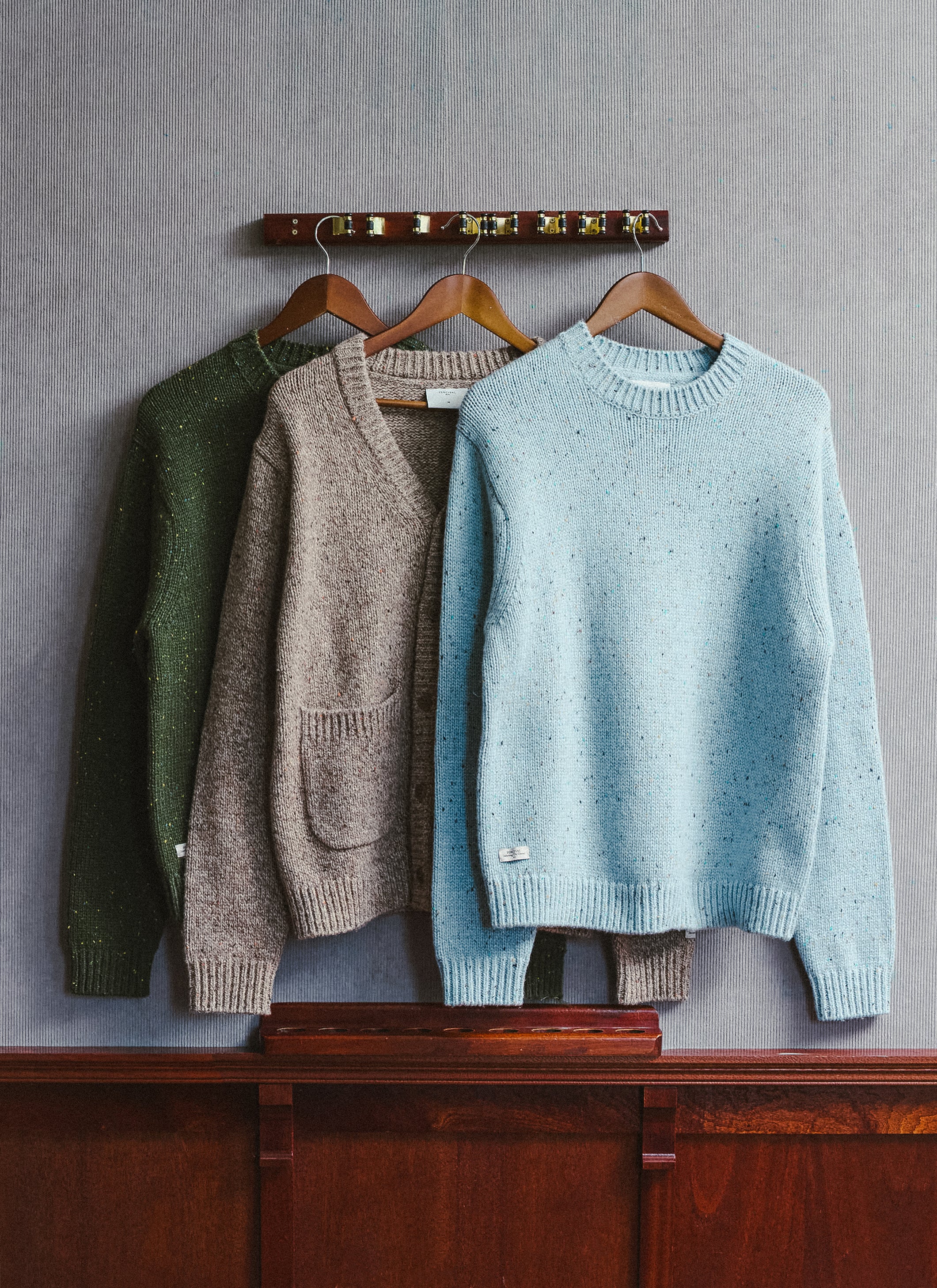 Three Percival knitwear pieces hanging on wooden hangers against a wall, showing green, brown, and blue colours.