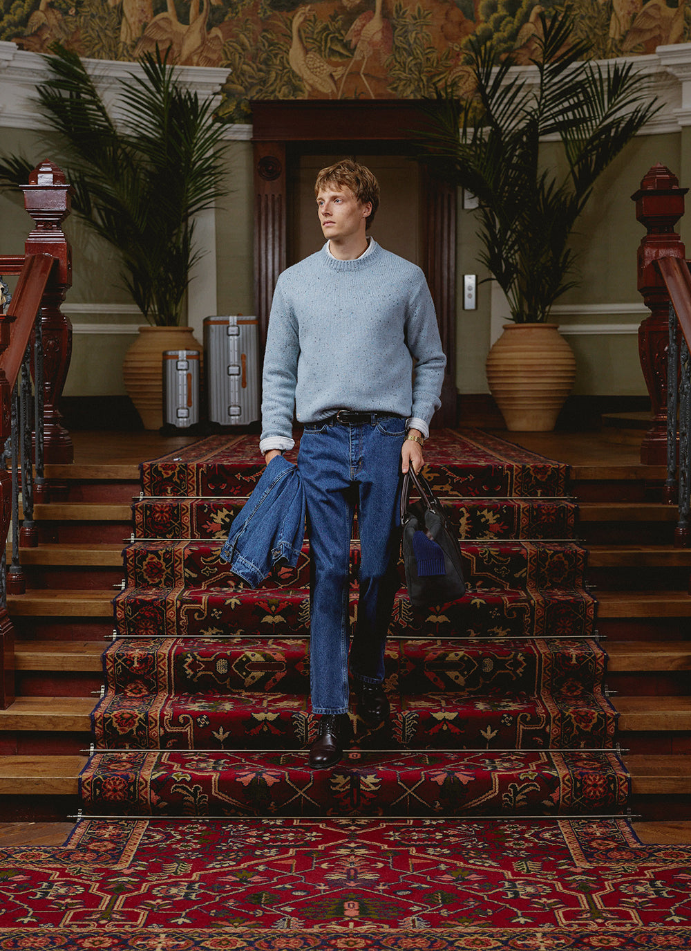 A model wears the blue Rhodes flecked jumper from Percival, styled with jeans and a holdall on a grand staircase.