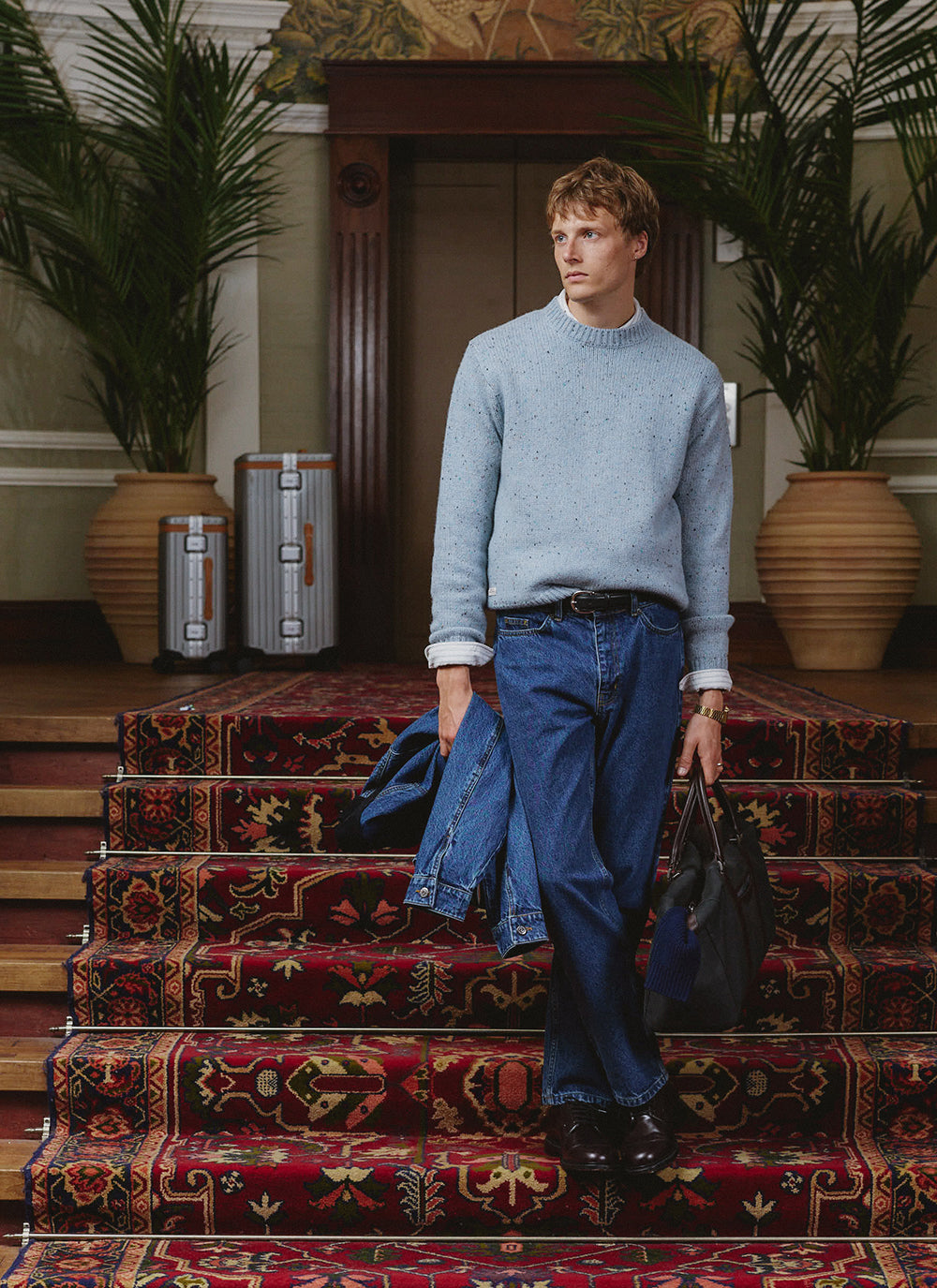 A model wears the blue Percival Rhodes crew neck jumper on a carpeted staircase, holding a denim jacket and bag.