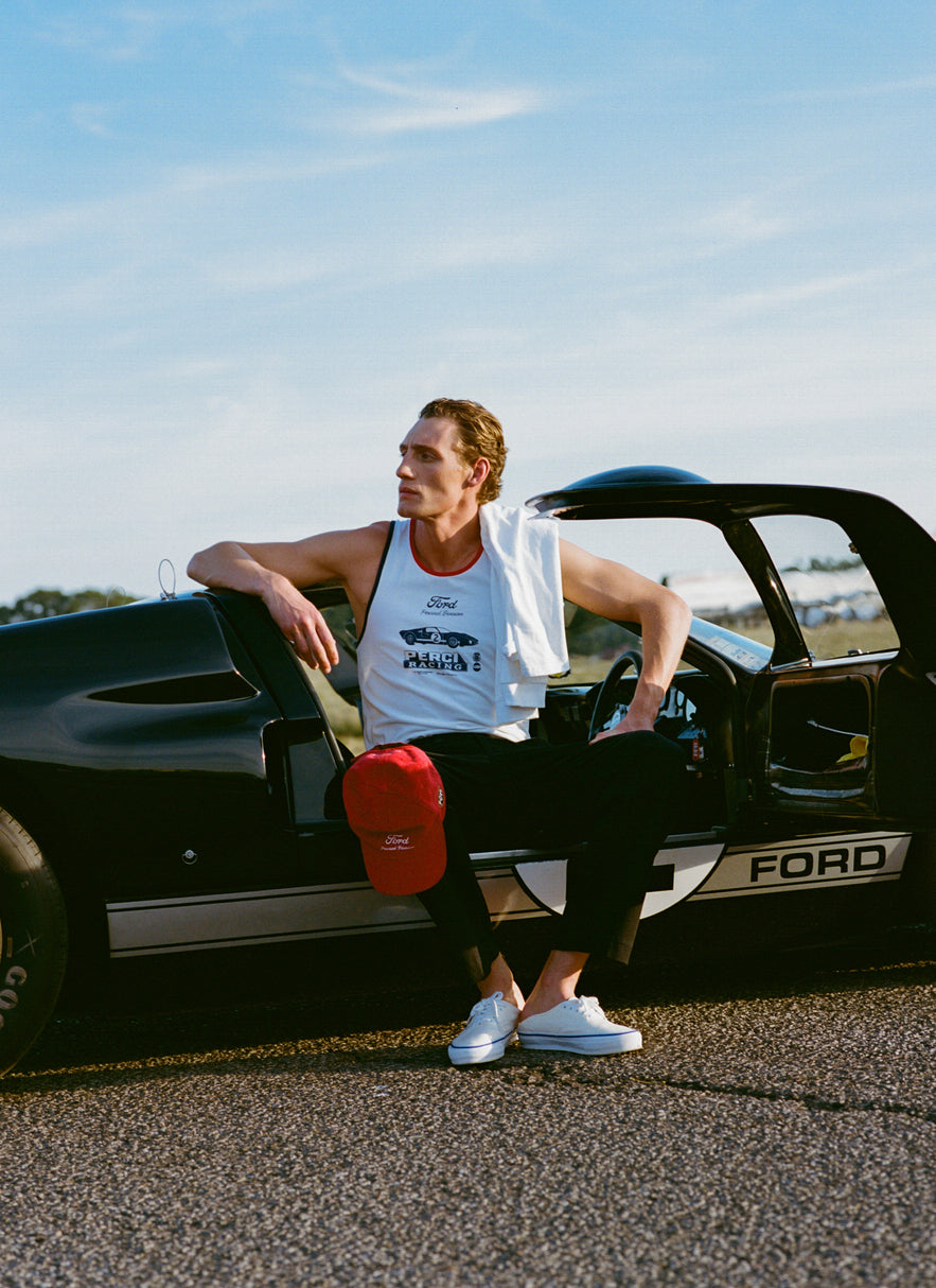 A model wears the white Percival x Ford GT40 ringer vest, sitting in a black Ford racing car with a red cap.