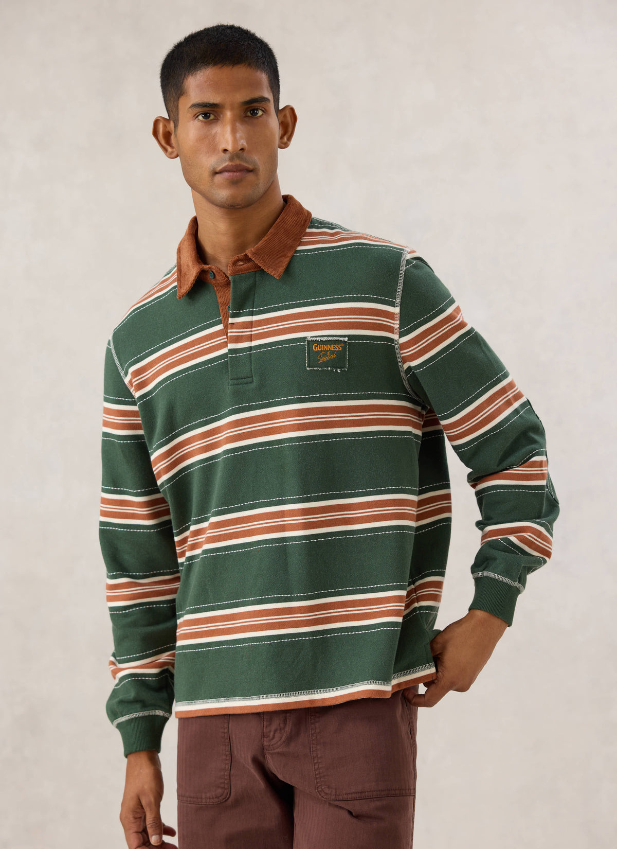 Model wearing the Guinness x Percival striped rugby polo in forest green with a brown cord collar and chest patch.
