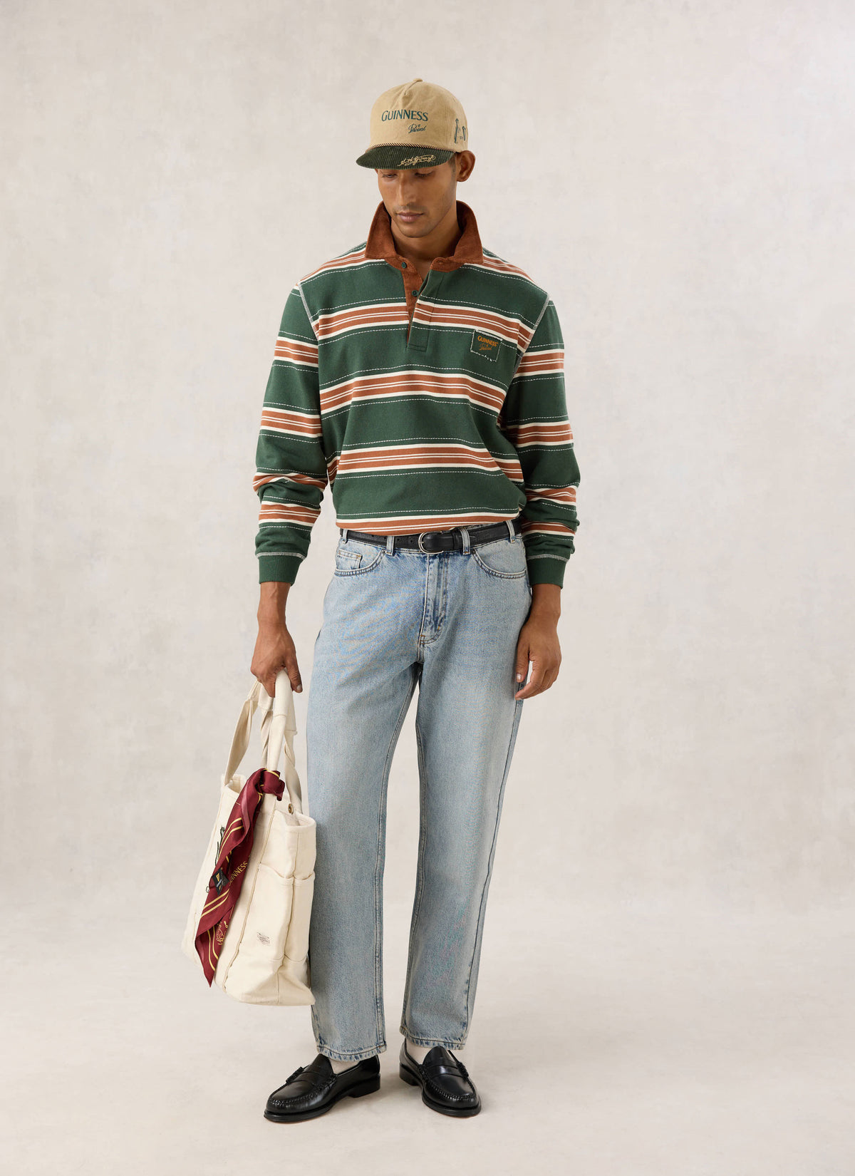 Model wearing the Guinness x Percival striped rugby polo shirt in forest green with a cord collar and cap.