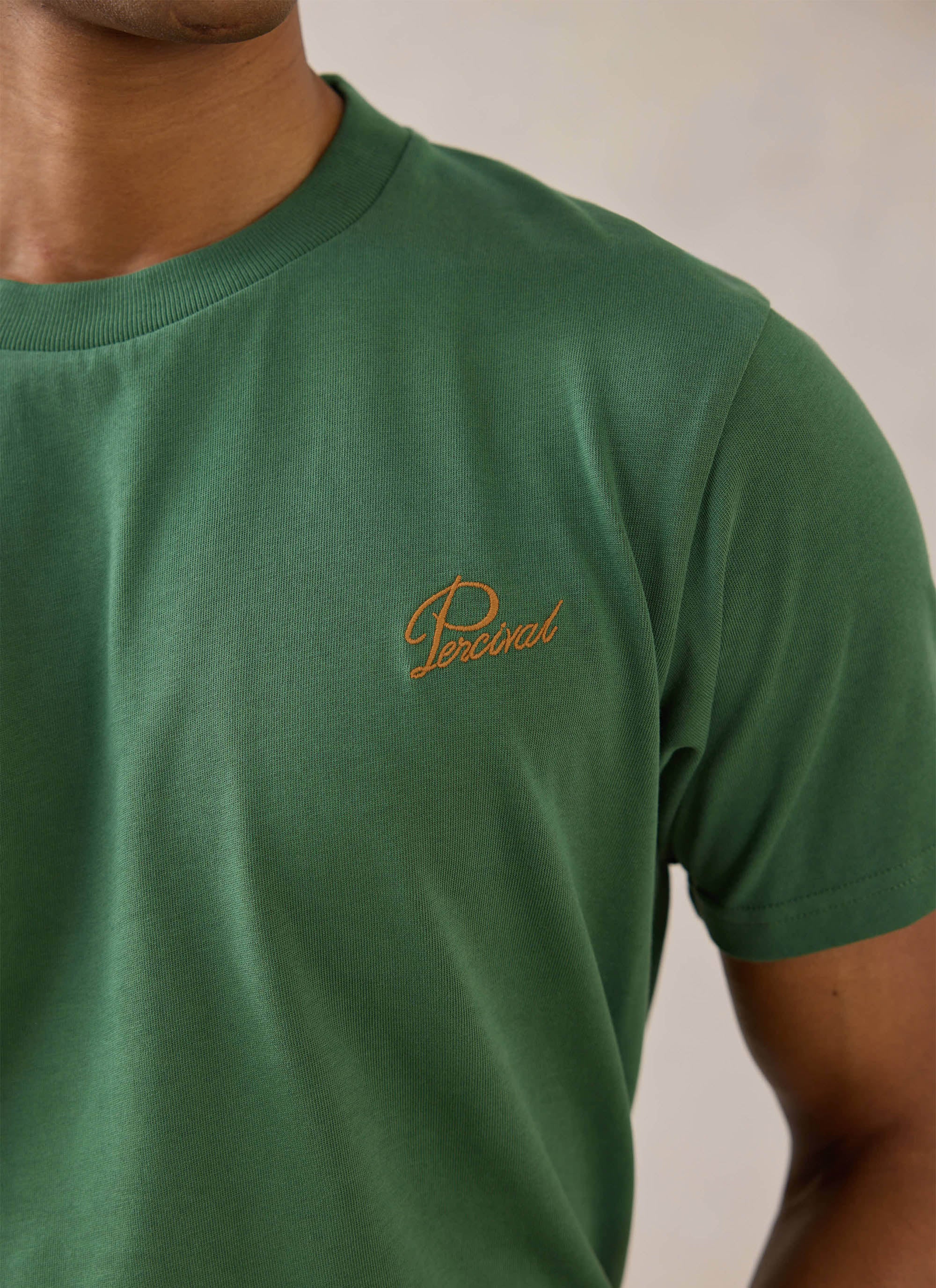 Close-up of a model wearing a forest green organic cotton Percival T-shirt with contrast script chest embroidery.