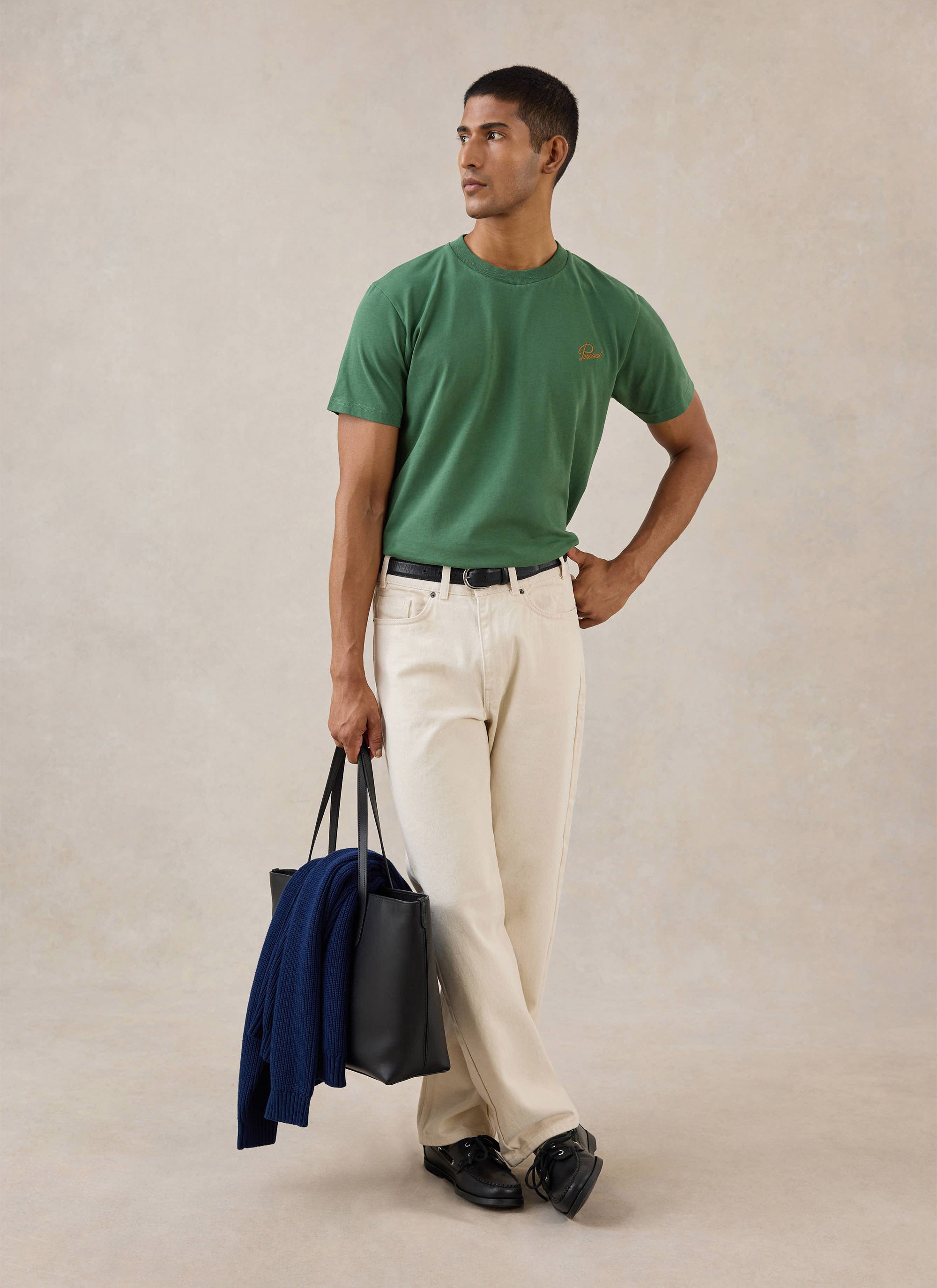 A model wears the forest green Percival Script Embroidered T-Shirt with cream trousers and a black tote bag.
