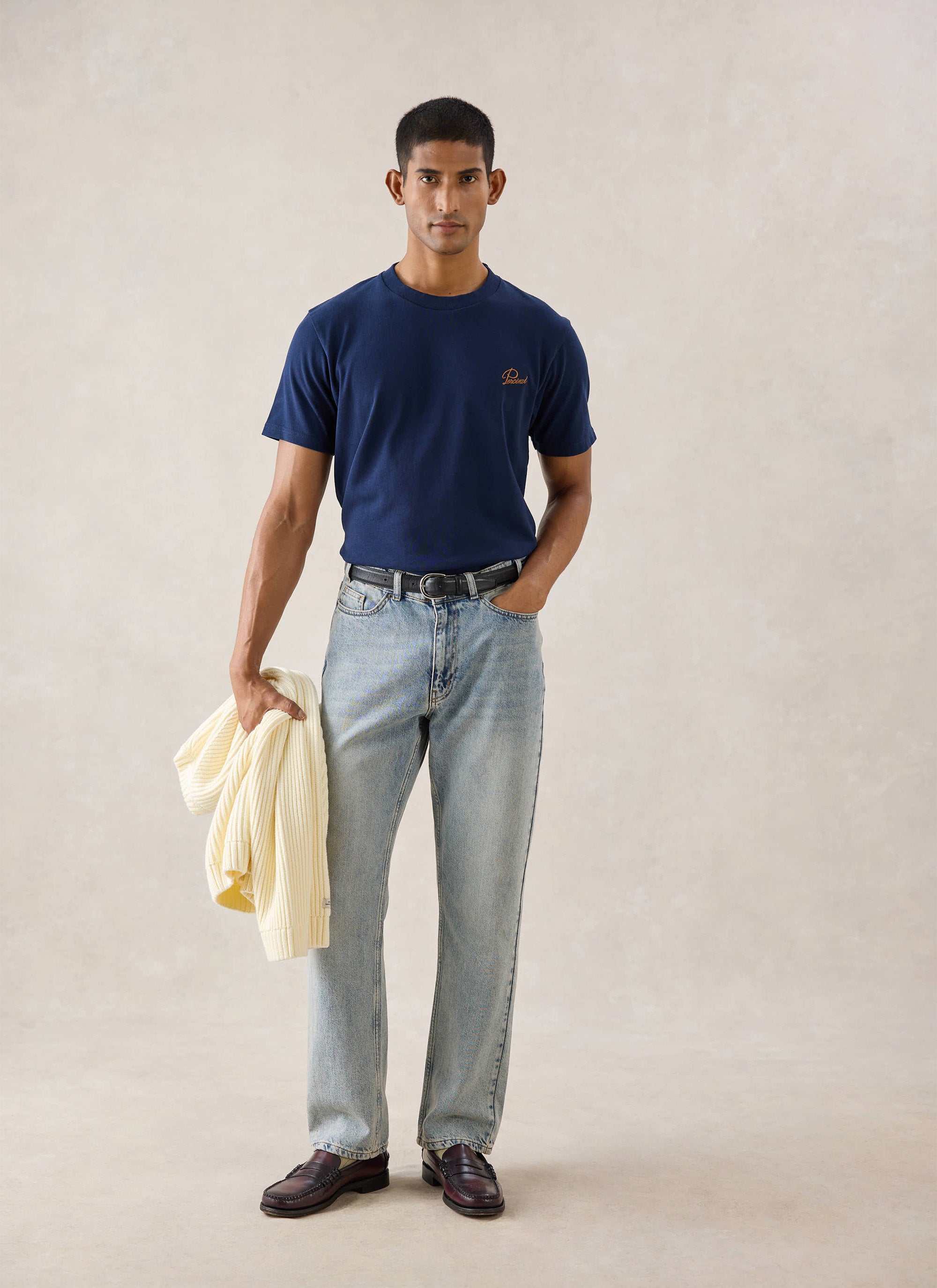 A model wears the navy Percival Script Embroidered T-Shirt with light wash jeans and loafers, holding a cream knit.