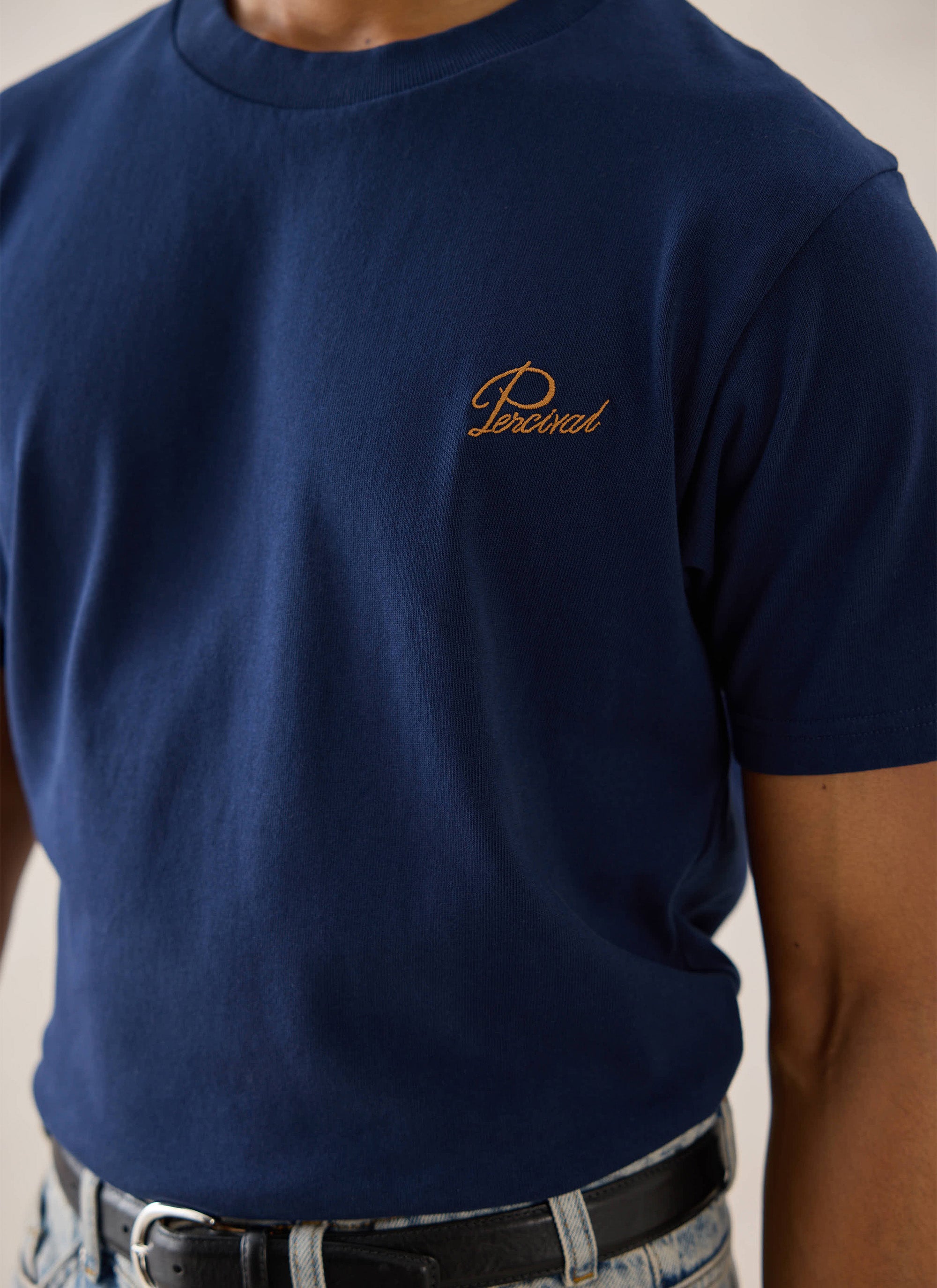 Close-up of a navy Percival organic cotton t-shirt featuring a small script embroidered logo on the chest.
