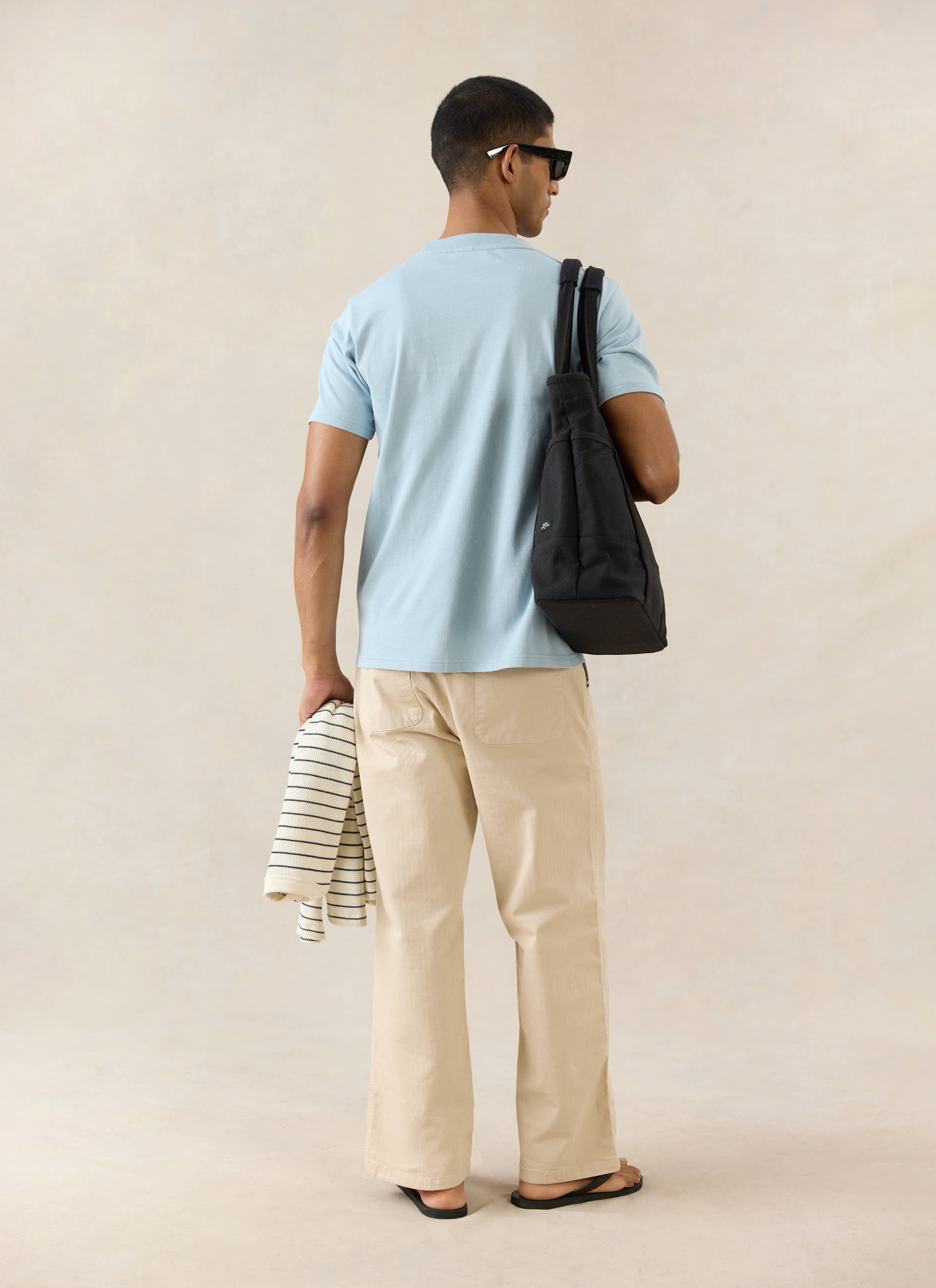 The Percival Perci Script T-shirt in light blue organic cotton, styled from behind with cream trousers and a black tote bag.