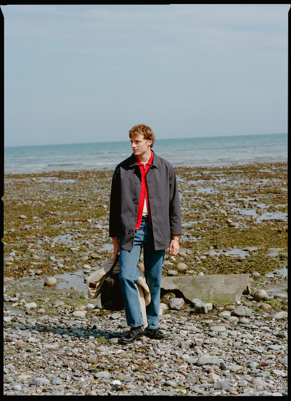 A model on a stony beach wearing a Percival waterproof short trench coat over a red zip-up top and blue jeans.