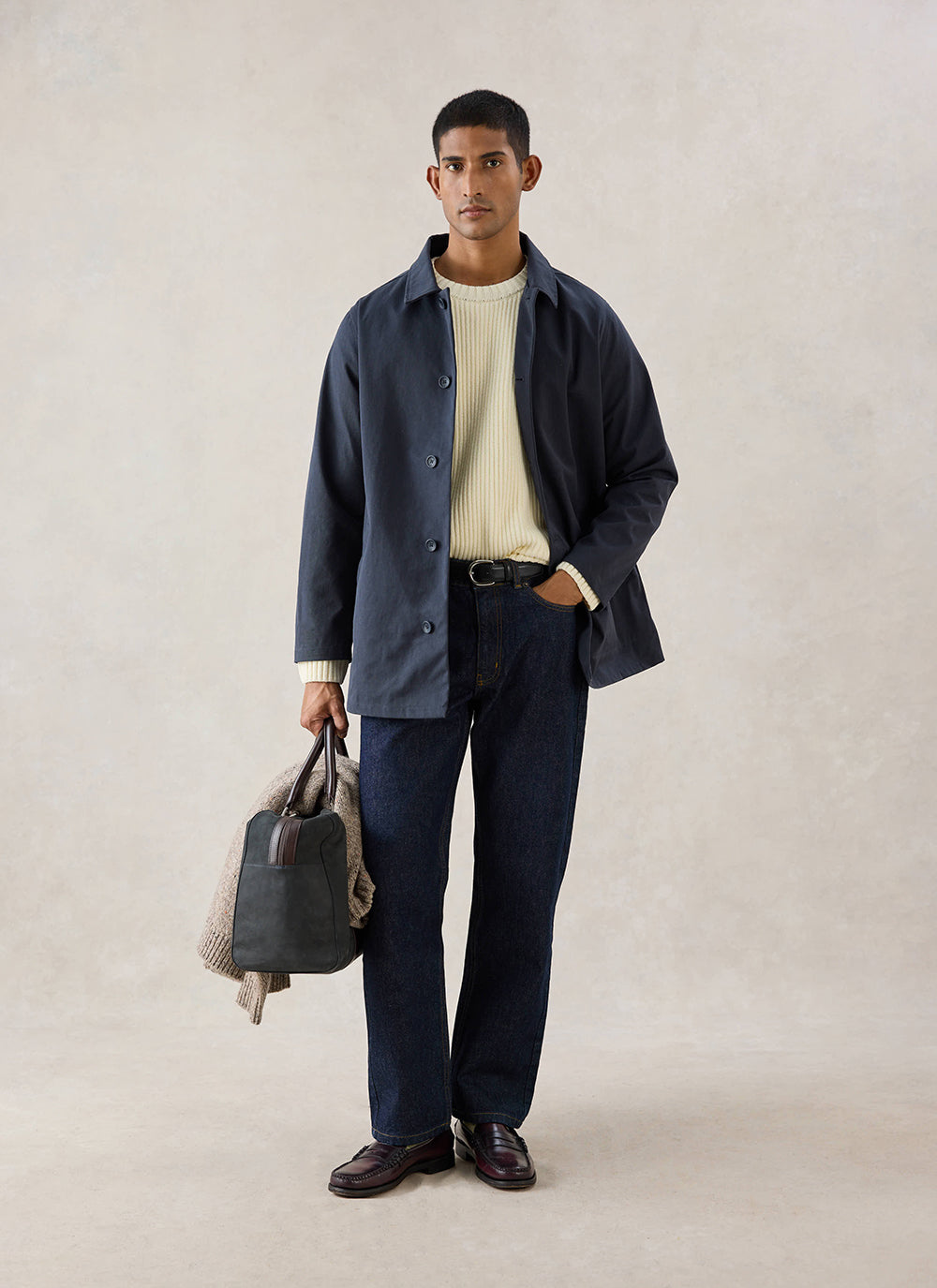 A model wears the navy Percival Sherlock short trench coat over a cream knit jumper, dark jeans and loafers, holding a bag.