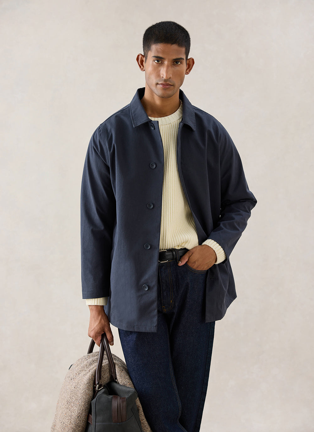 A model wears the navy Percival Sherlock short waterproof trench coat over a cream knit jumper and dark jeans, holding a bag.