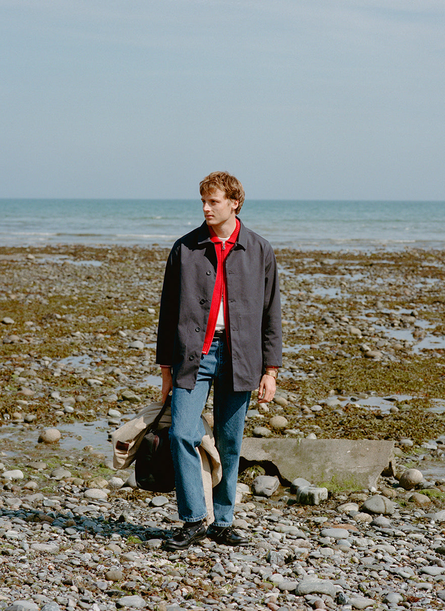 A model on a beach wearing a Percival navy trench coat over a red zip cardigan and denim jeans.