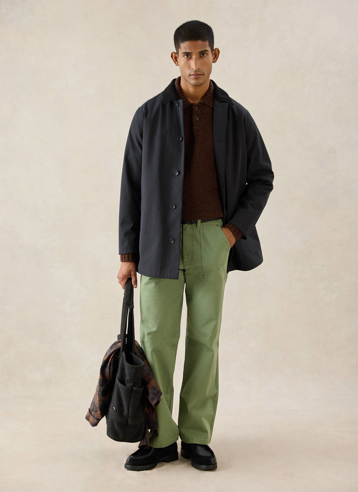 A model wears the black Percival trench coat, styled with green trousers, a brown knit jumper, and black shoes.