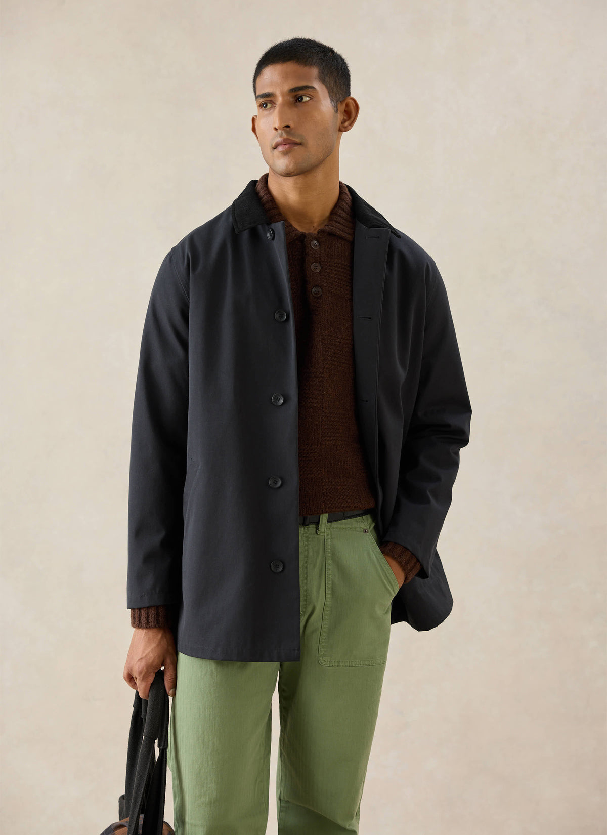 A model wears the black Percival Sherlock short trench coat, styled with a brown knit polo and green trousers.