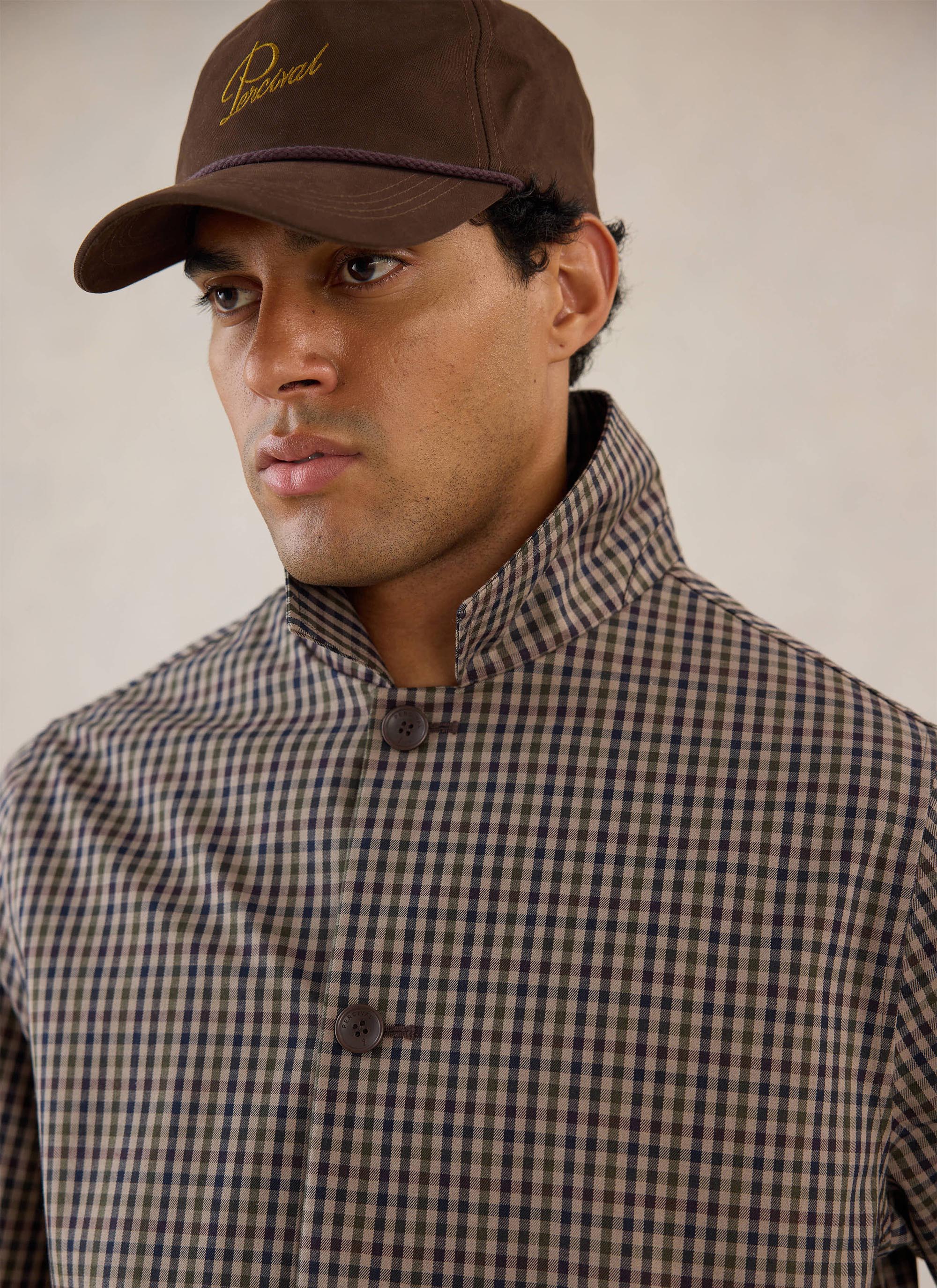 Close-up of a model wearing the Watcher Check Percival Sherlock Short Trench Coat with high collar and brown cap.