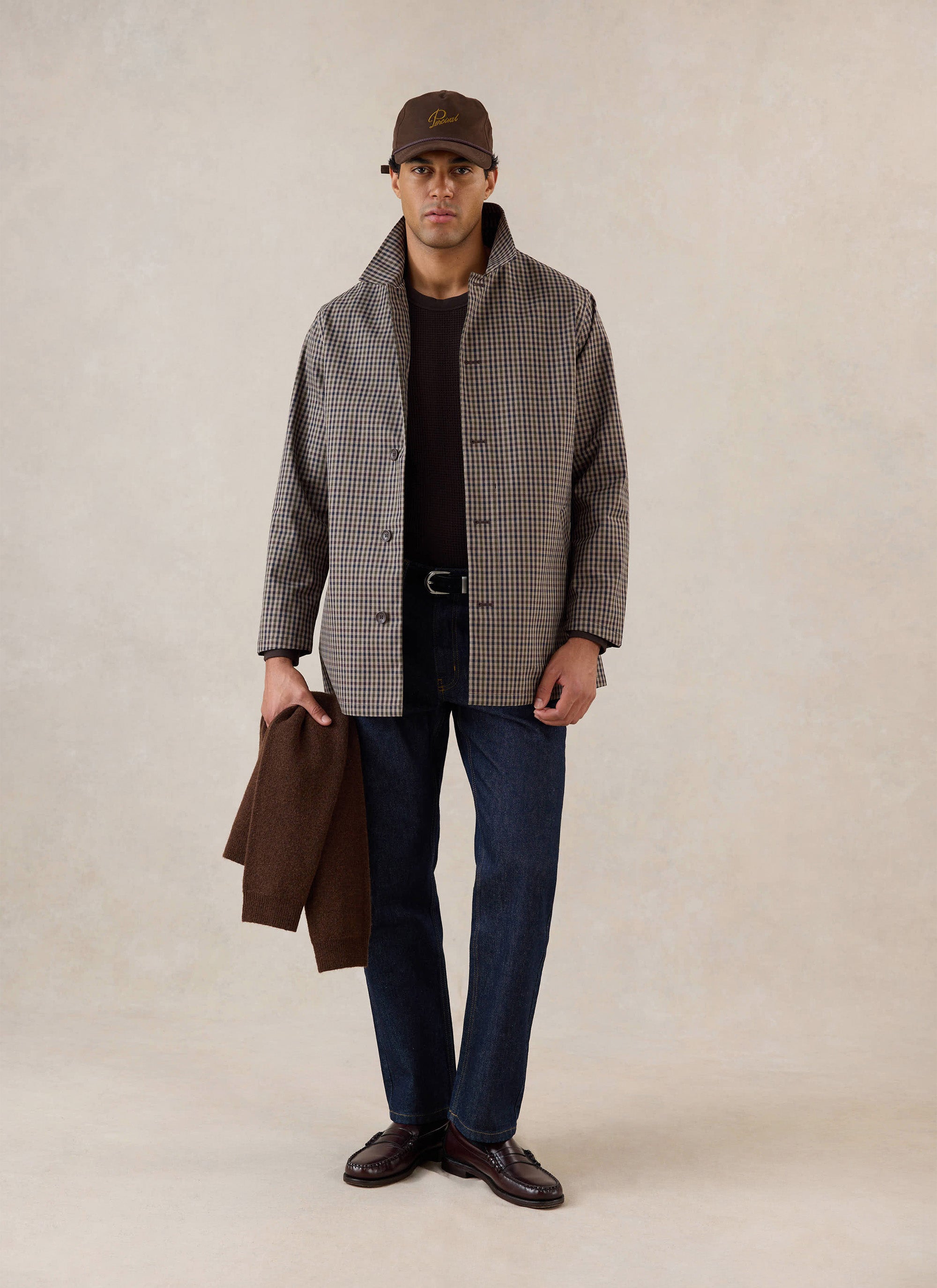 A model wears the Percival Sherlock short trench coat in Watcher Check, dark jeans, and loafers in a studio.