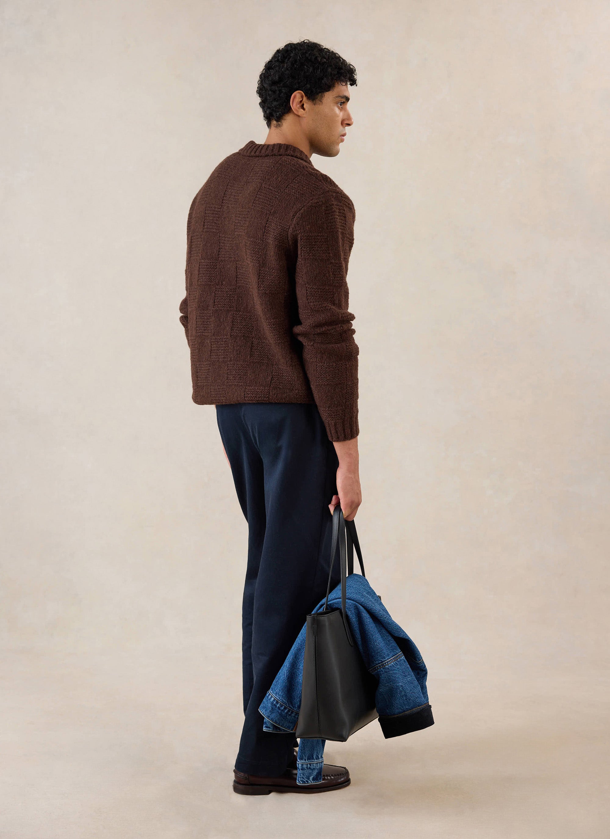 A model wears the brown Percival Shugart boxy knit jumper, holding a black tote bag and denim jacket in a studio.