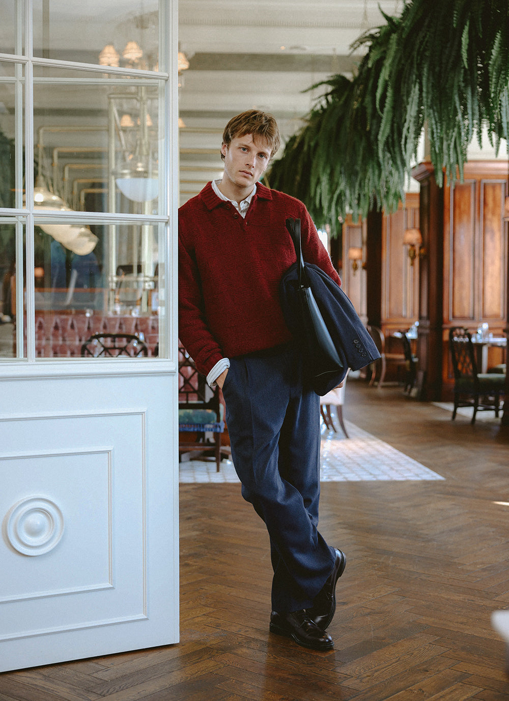 A model wears the burgundy Percival Shugart knit jumper with trousers in a restaurant setting.