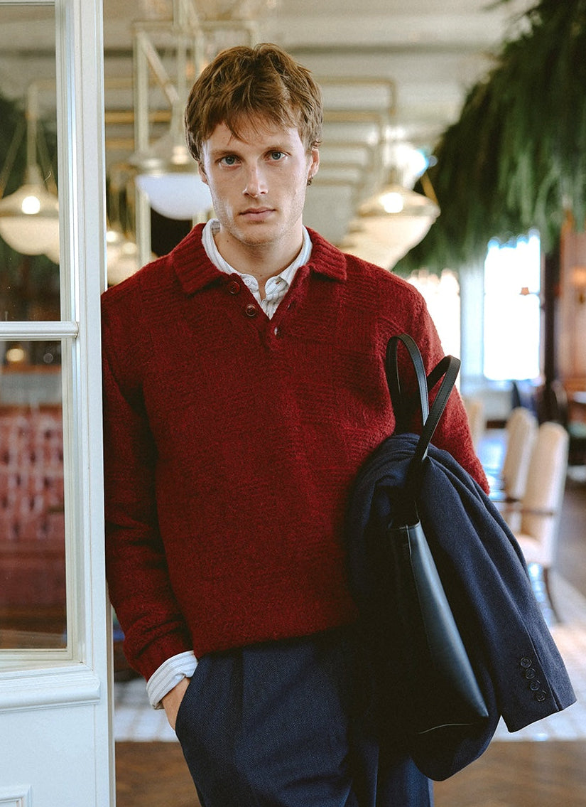 A model wears the burgundy Percival alpaca wool Shugart knit jumper over a collared shirt, holding a bag and jacket.