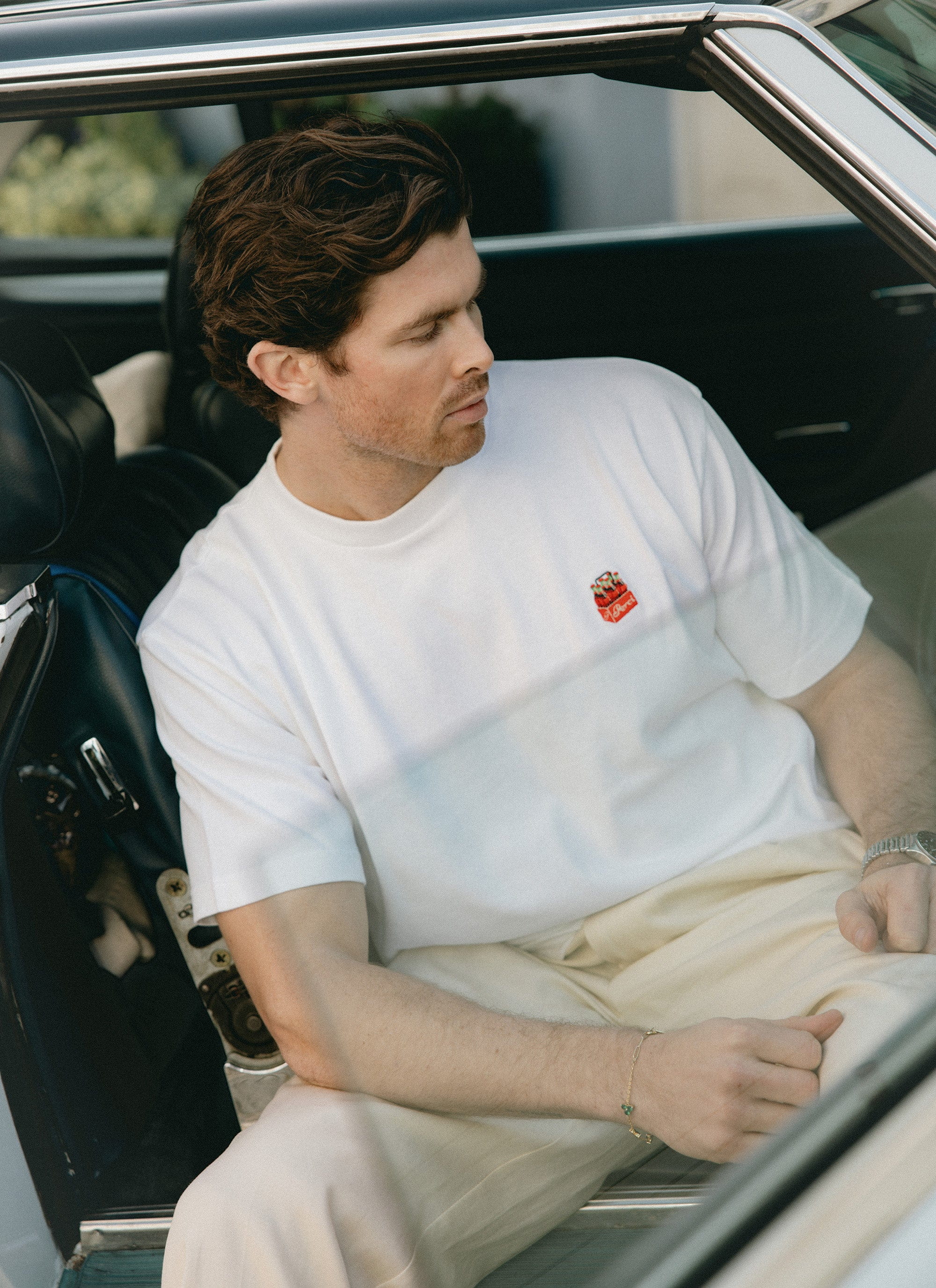 Lifestyle shot of a model wearing the white Percival Soda Bottles Oversized Embroidered T Shirt in a vintage car.