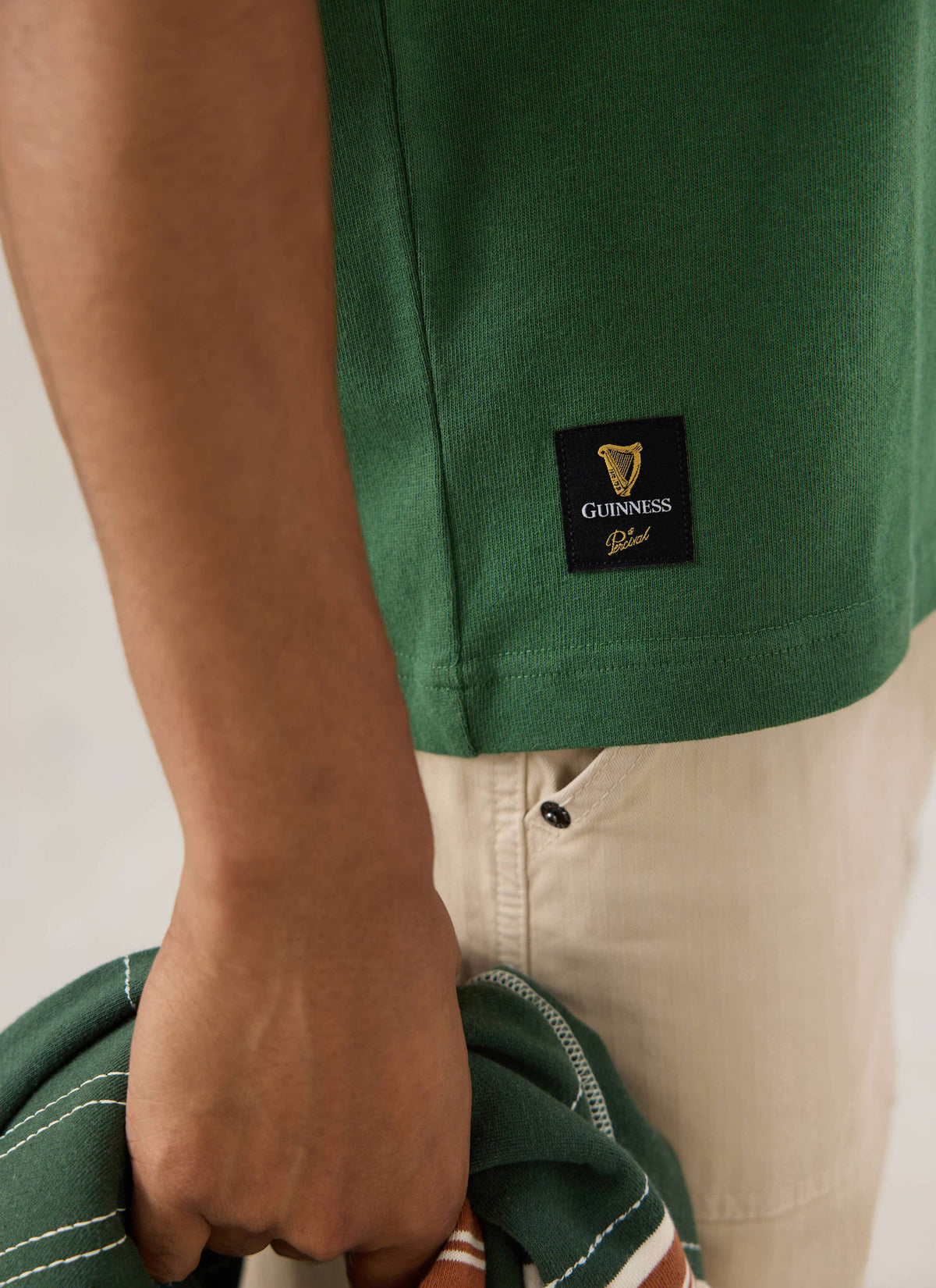 A close-up of the forest green Percival x Guinness t-shirt showing the branded hem tab and heavyweight cotton texture.