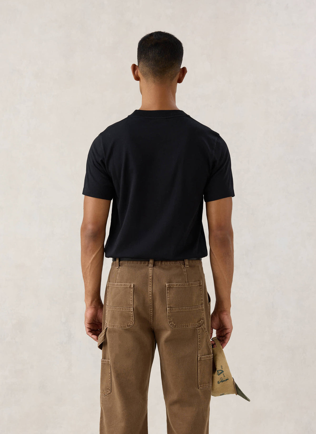 Rear view of a model wearing the black Guinness x Percival Stout T-Shirt and brown utility trousers.