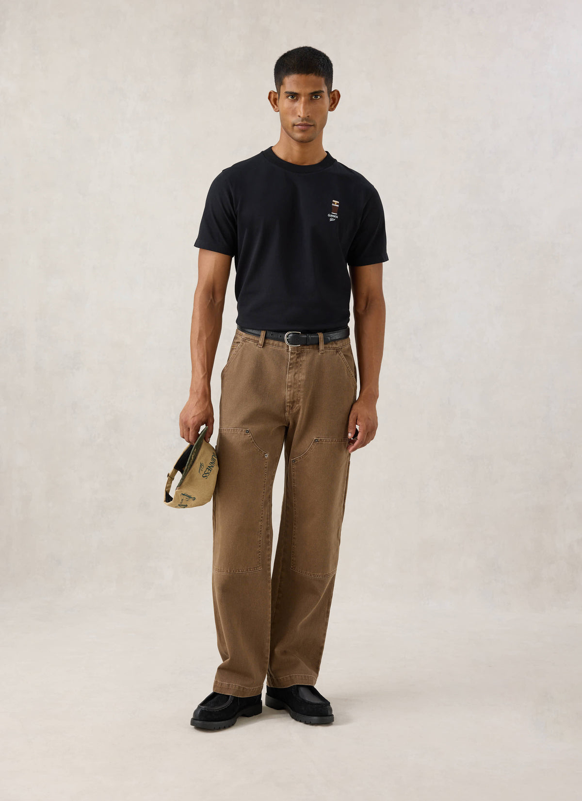 Model wearing the black Guinness x Percival stout t-shirt with chest embroidery, paired with brown trousers.