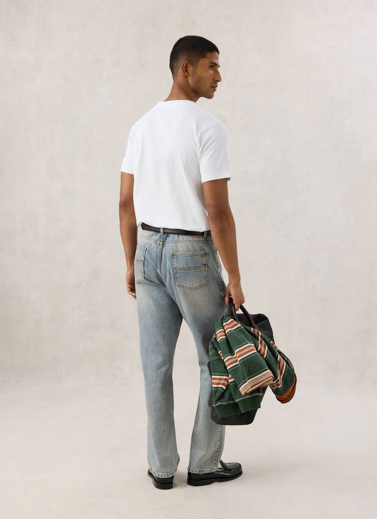 Rear view of a model wearing the white Guinness x Percival Stout T-shirt with light wash jeans and carrying a bag and jacket.