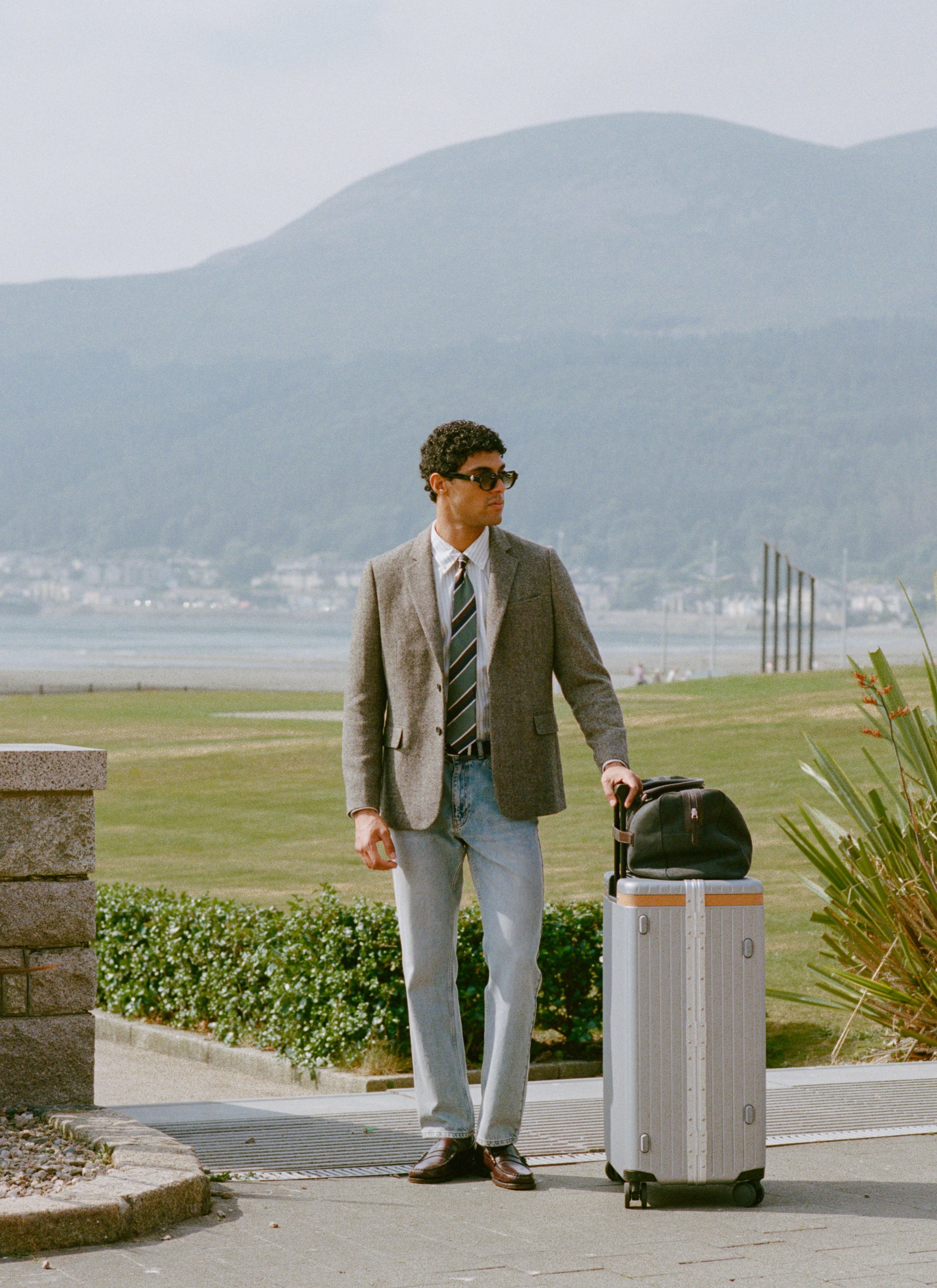 A model wears the Percival Tailored Blazer in Birdseye Wool Grey and Percival Stripe Tie in Silk Multi with a suitcase.