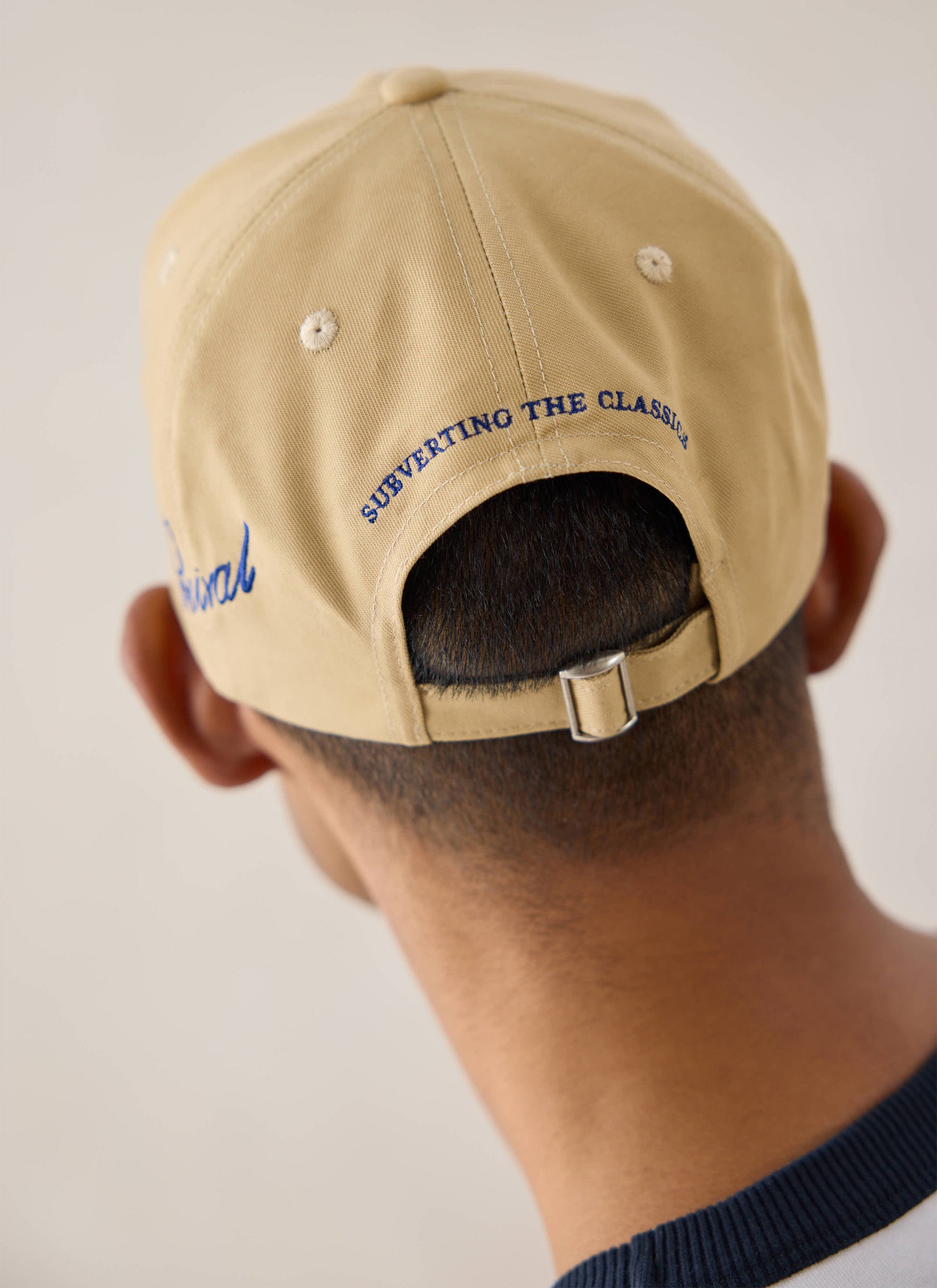 A close-up view of the back of the tan Percival trucker cap showing blue embroidery and strap closure.