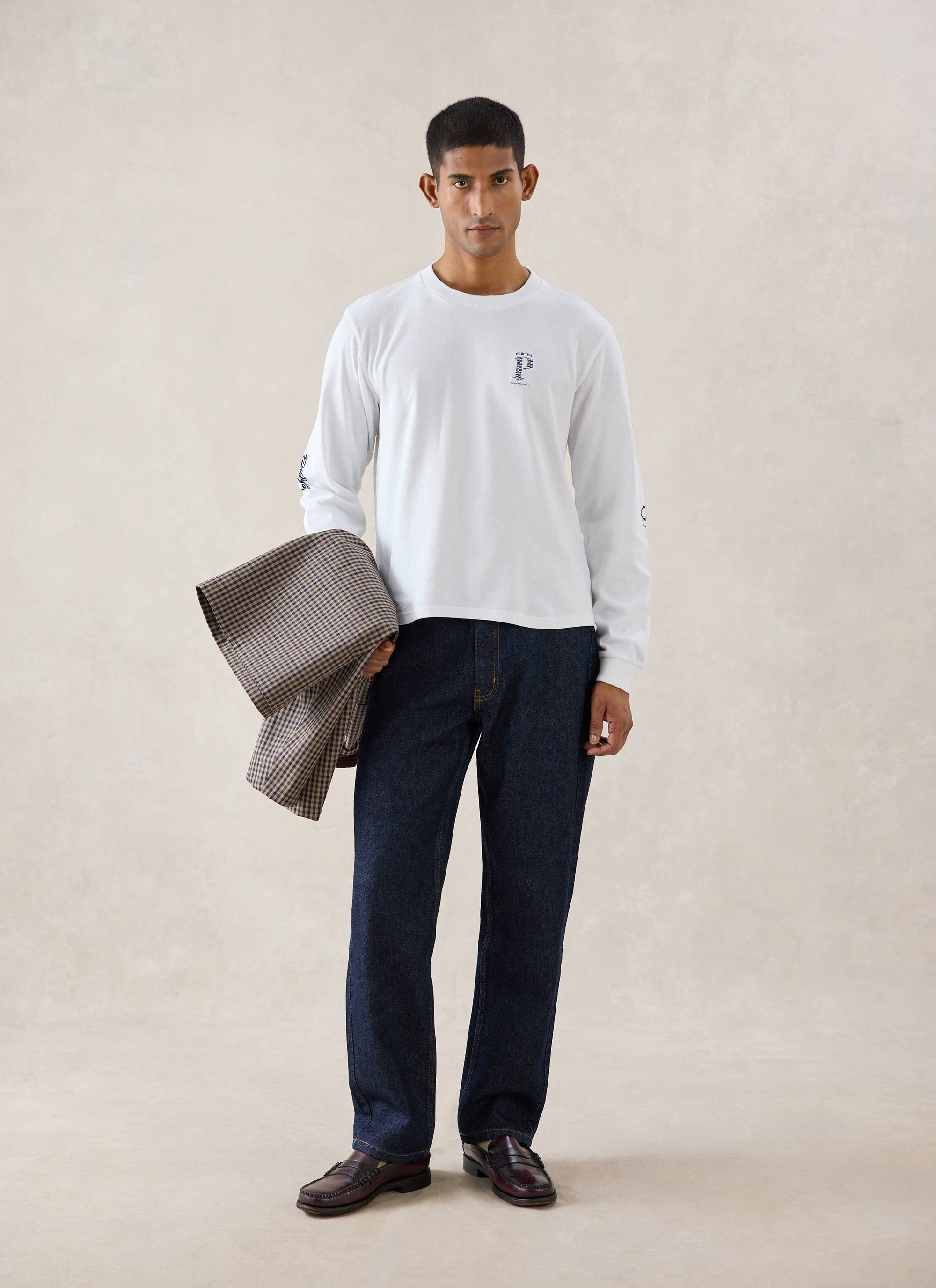 A model wears the white Percival Tucker long sleeve t-shirt and dark denim jeans, holding a checked jacket in a studio.