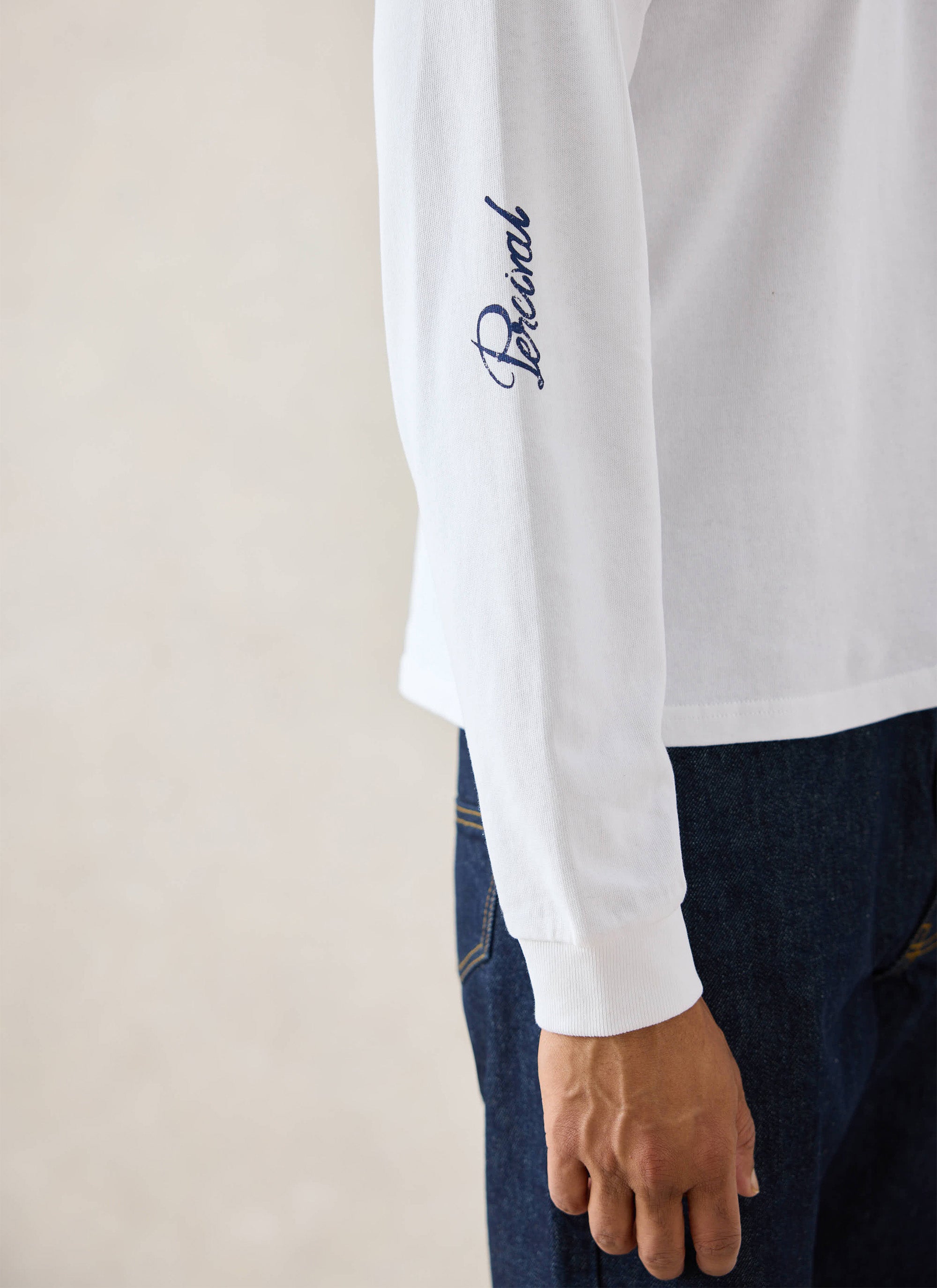 A close-up of a model wearing the white Percival Tucker long sleeve t-shirt, highlighting the blue sleeve embroidery.