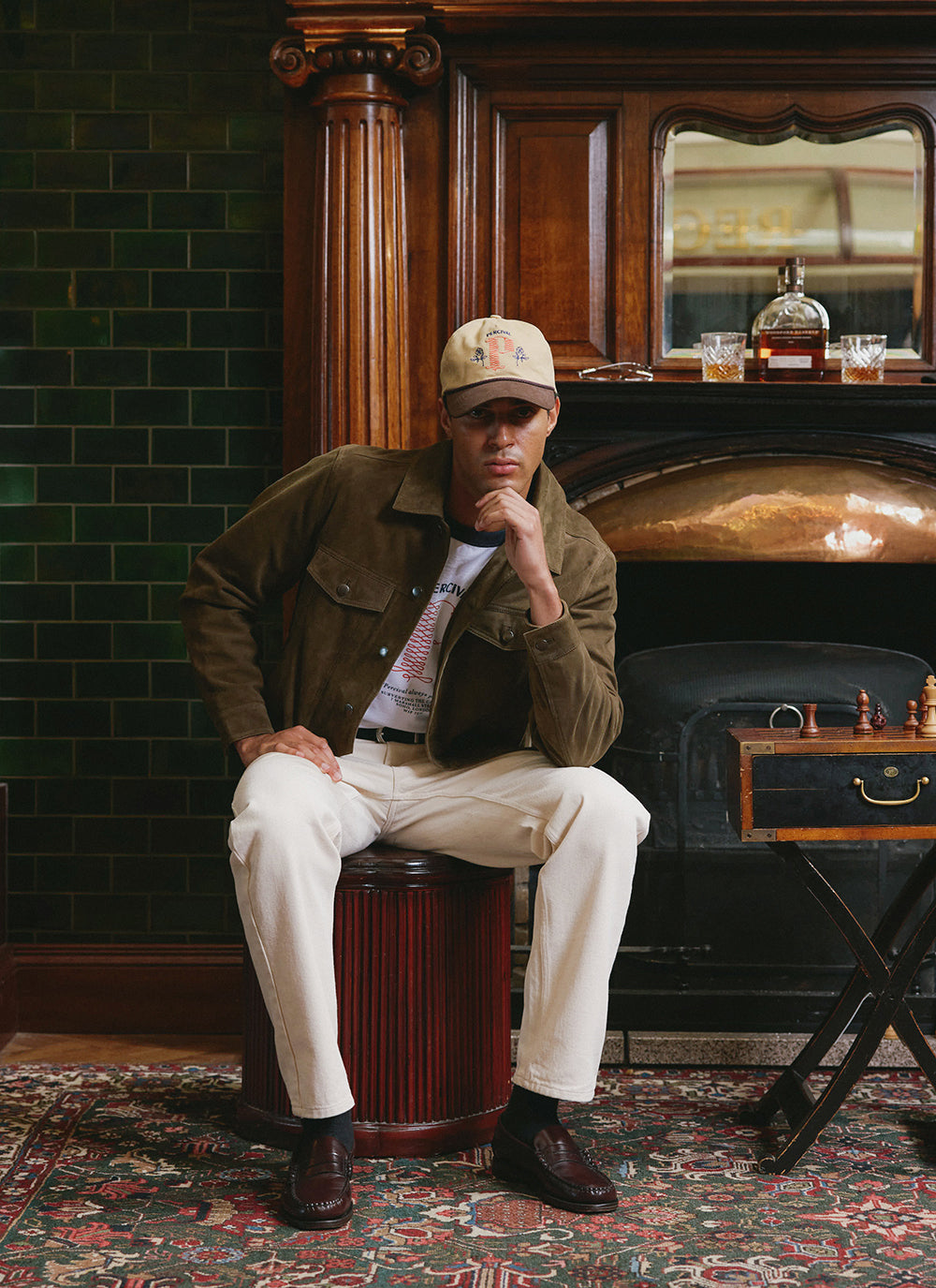 A model wearing a Percival olive suede western jacket with a graphic t-shirt, cream jeans, and a cap.