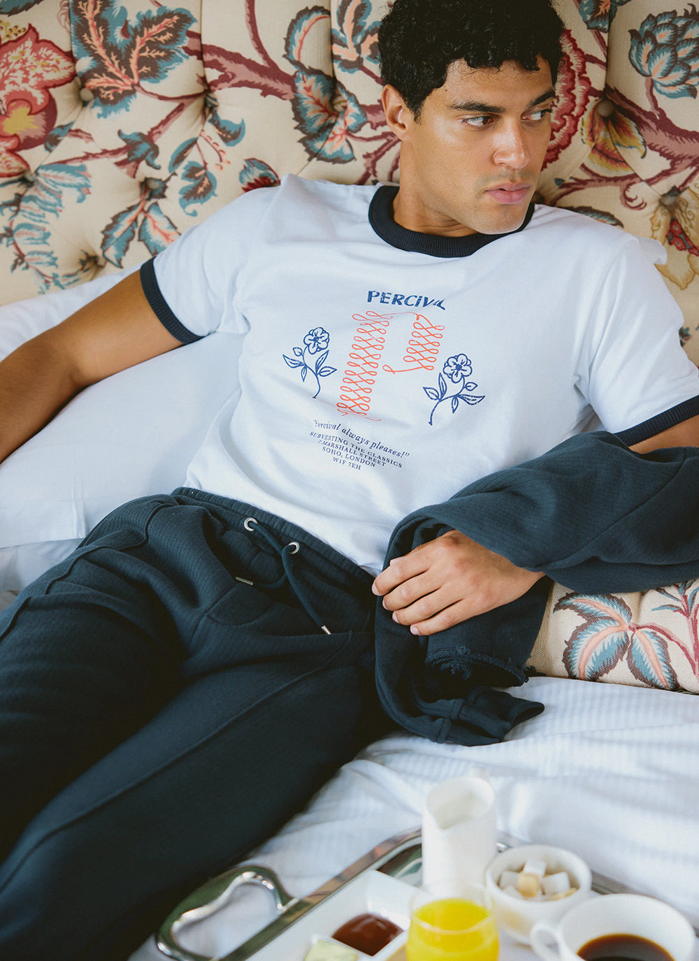 A model wearing Percival navy Conrad trackpants and a white ringer t-shirt while relaxing on a bed.