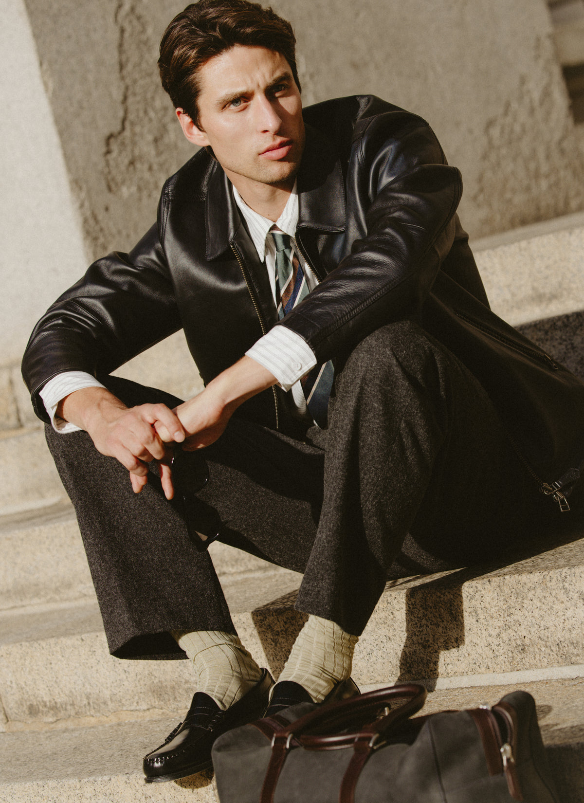 A model sitting outdoors wearing the black Percival Turner leather zip jacket over a striped shirt and charcoal trousers.