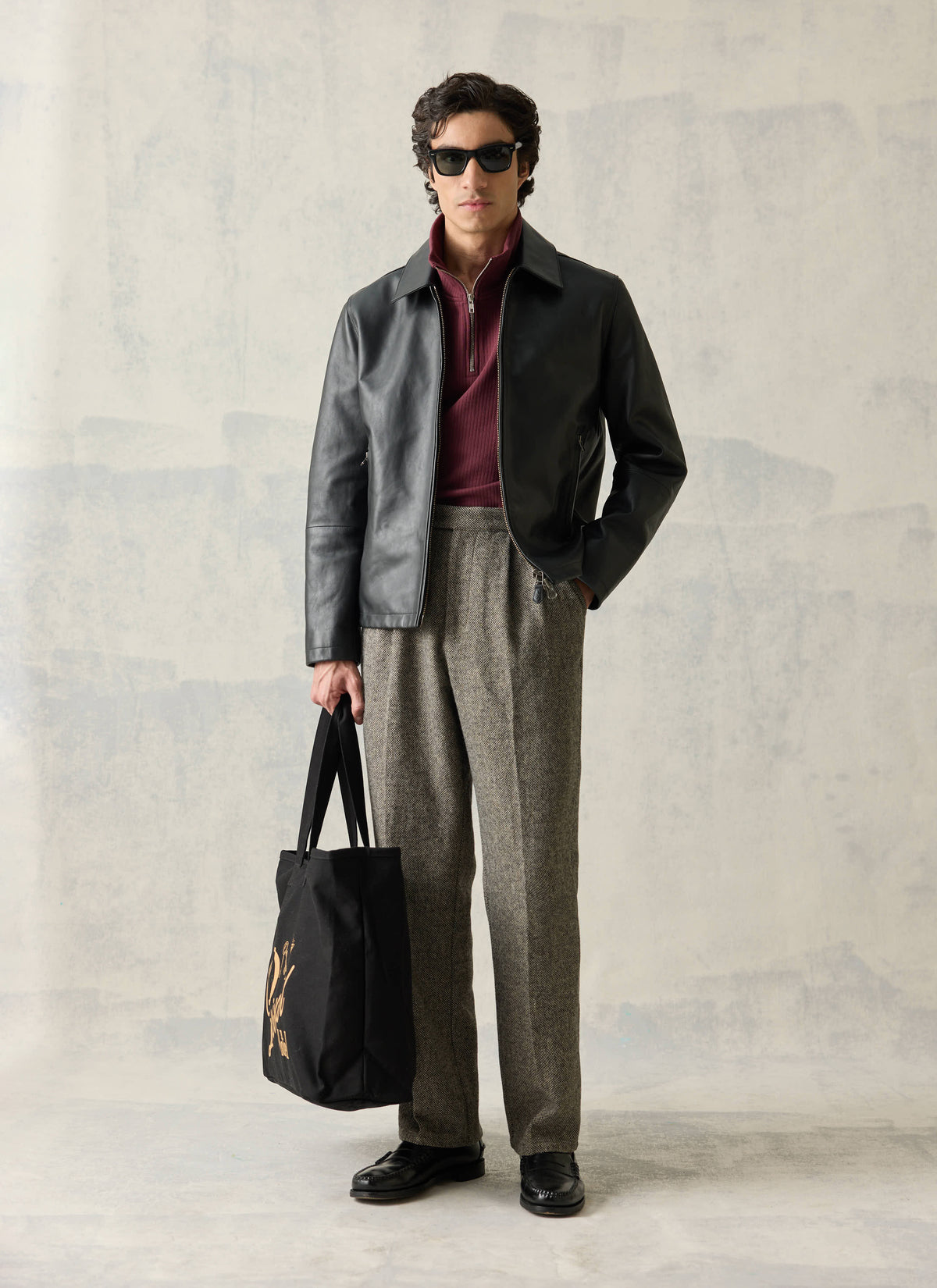 Model wearing the black Percival Turner leather zip jacket and trousers while holding a black tote bag.
