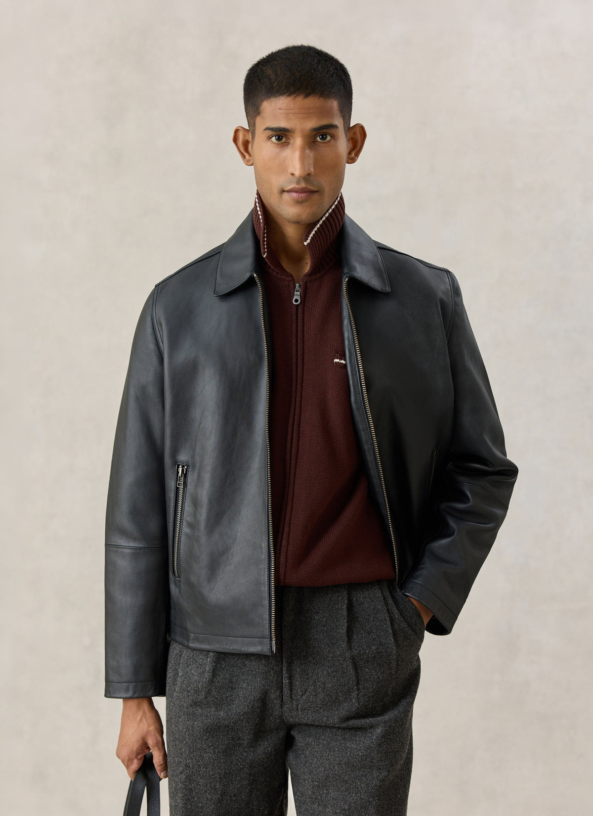 A model wearing the Percival black leather Turner zip jacket over a dark red knit zip-up and grey trousers.