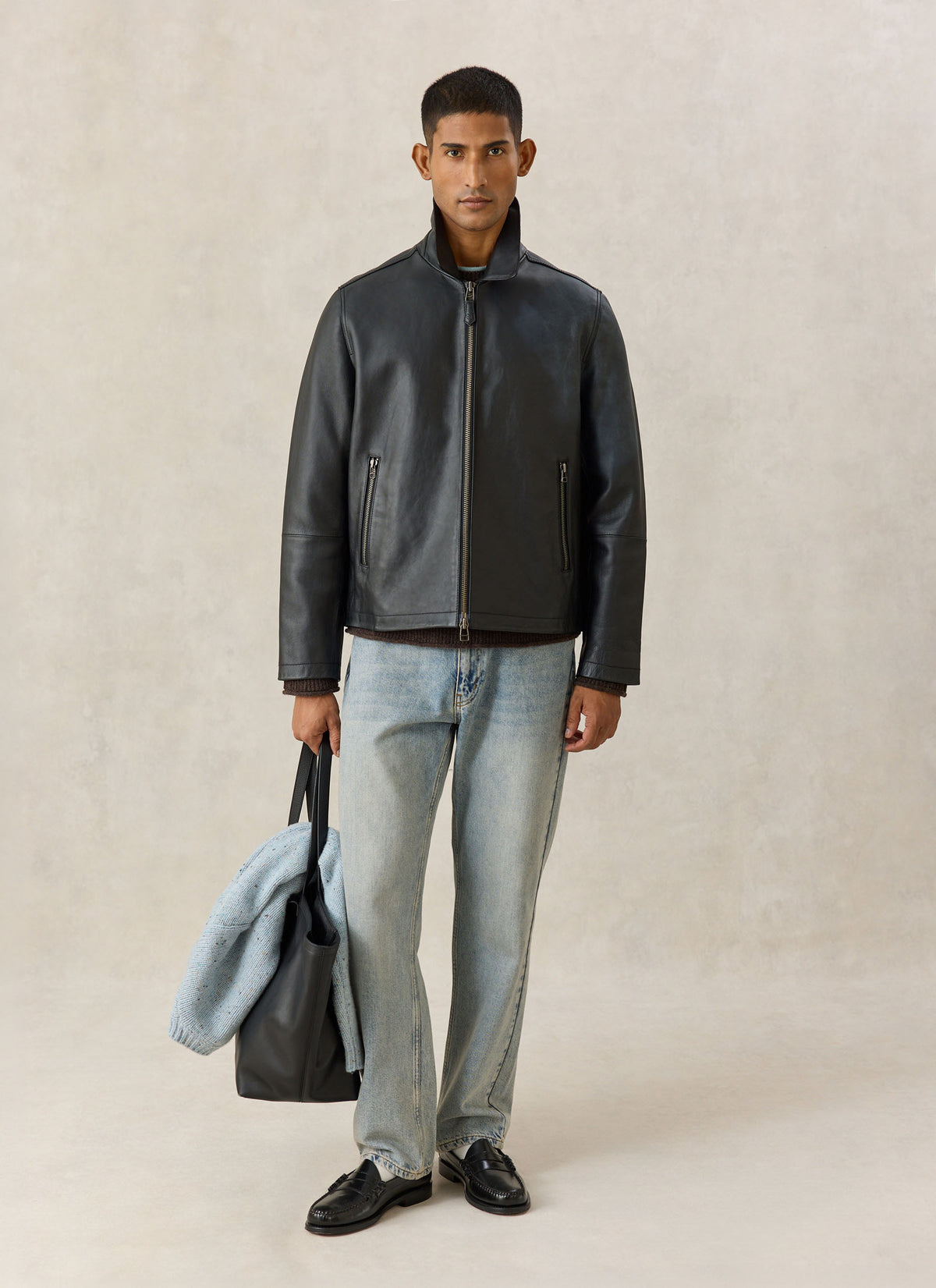A model wearing the Percival Turner black leather zip jacket with light wash jeans, holding a black leather tote bag.
