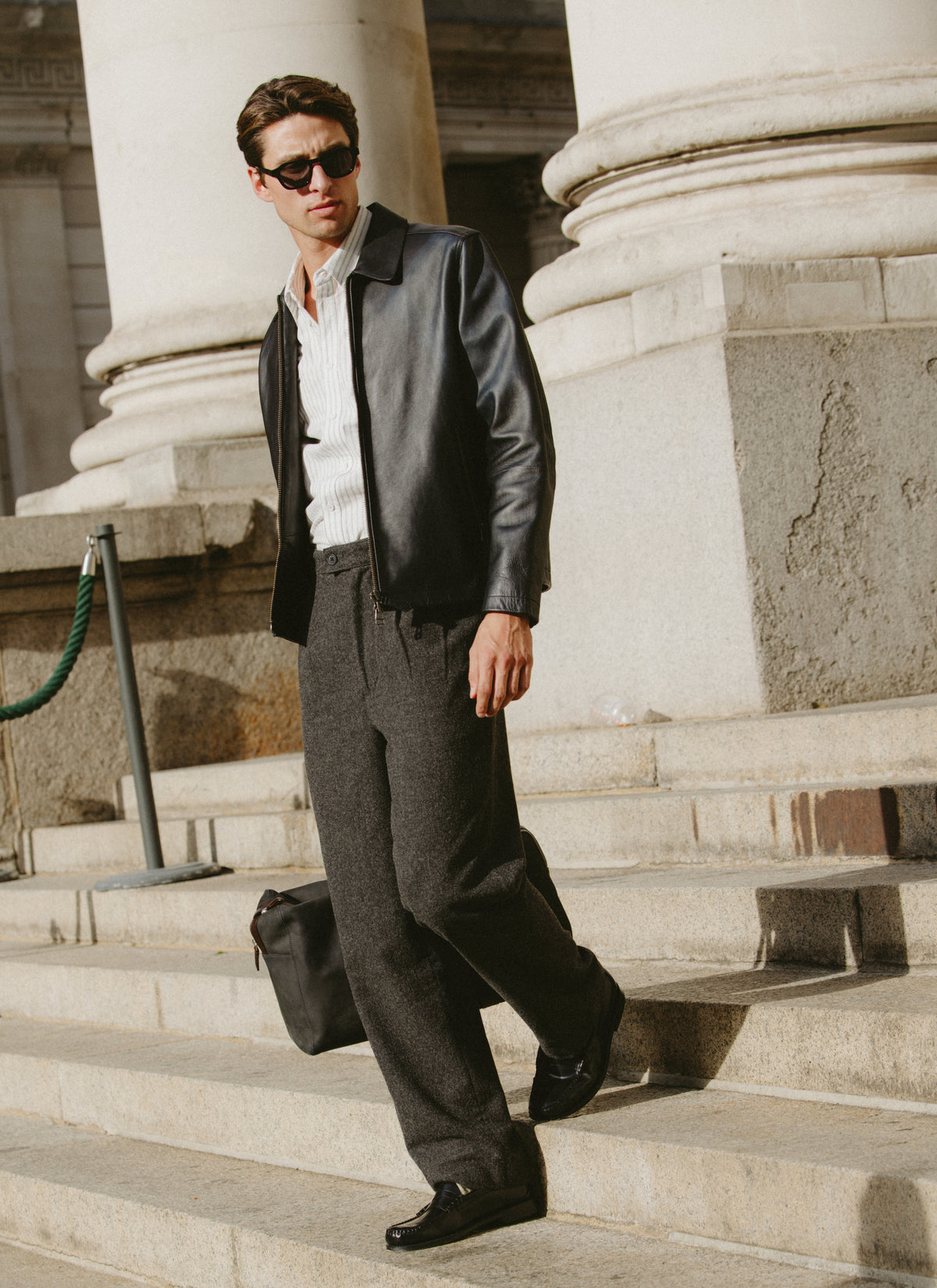 A model wearing the black Percival Turner leather jacket and charcoal Blake trousers while walking down stone steps outdoors.
