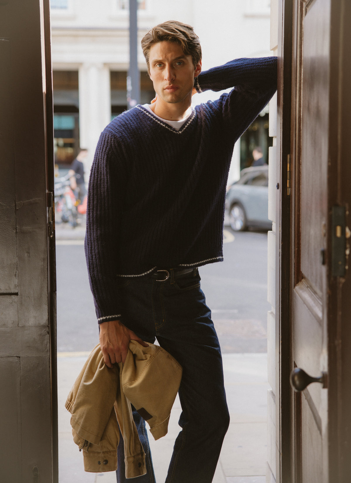 A model wearing the navy Percival Vincent V-neck jumper, holding a jacket while standing in a doorway.