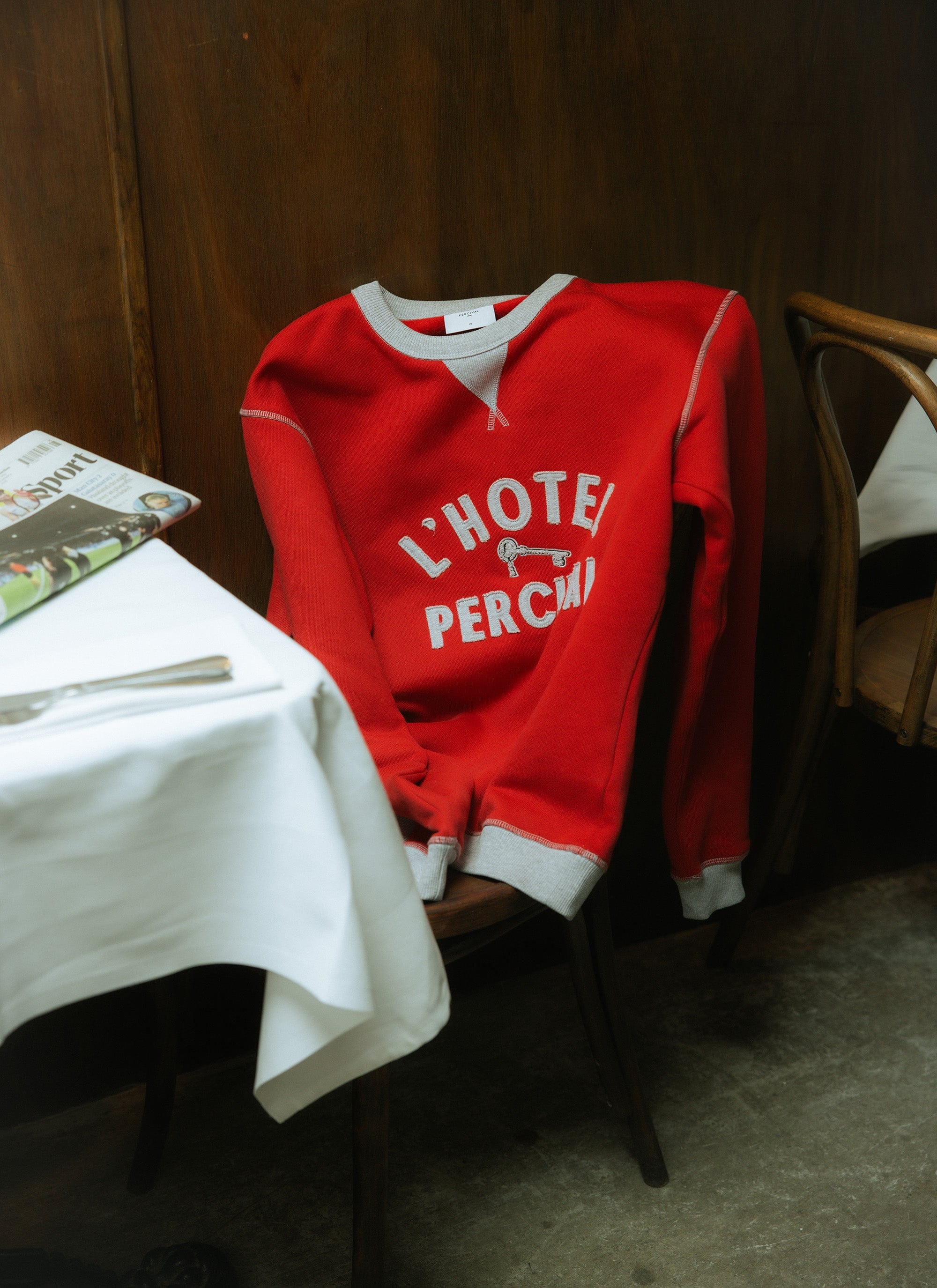 The Percival Ronald Sweatshirt in red organic cotton, with 'L'Hotel' appliqué lettering, casually draped over a wooden chair.