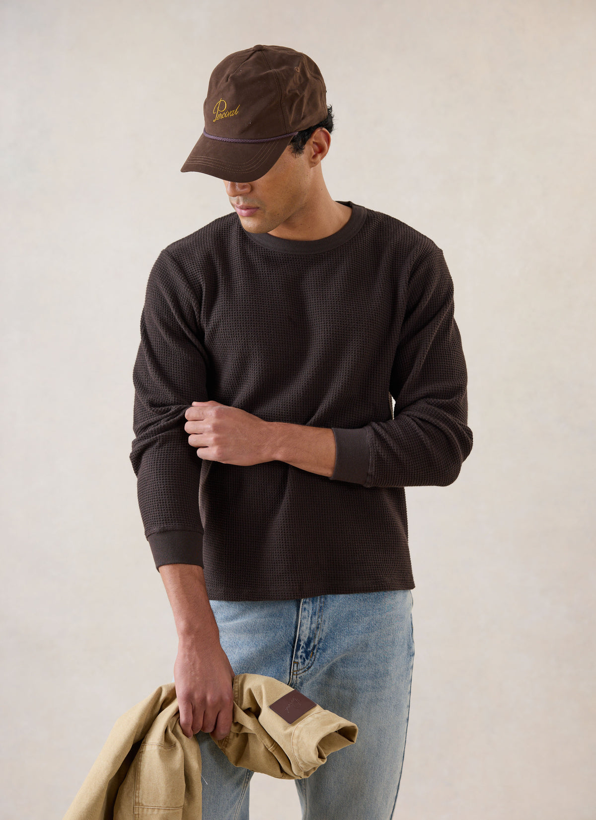 A model wearing a brown Percival waffle long-sleeve T-shirt, Luther cap, while holding the Jackson jacket.